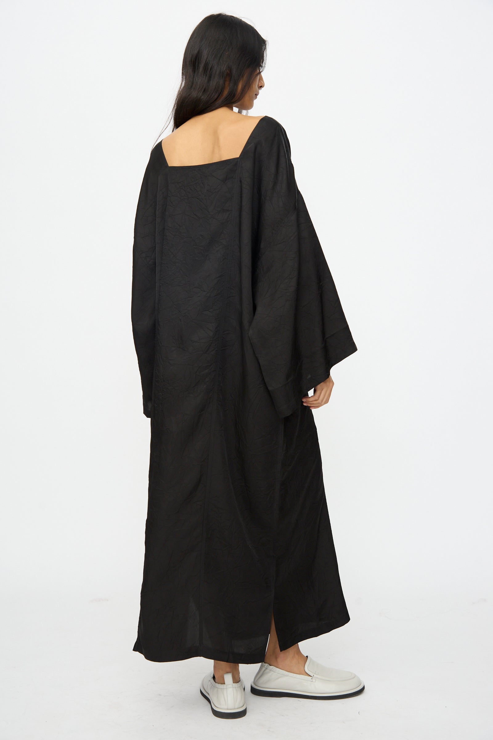 A person wearing the Cristaseya Square Neck Caftan Dress in Black with wide sleeves and white shoes stands facing away from the camera against a plain white background.