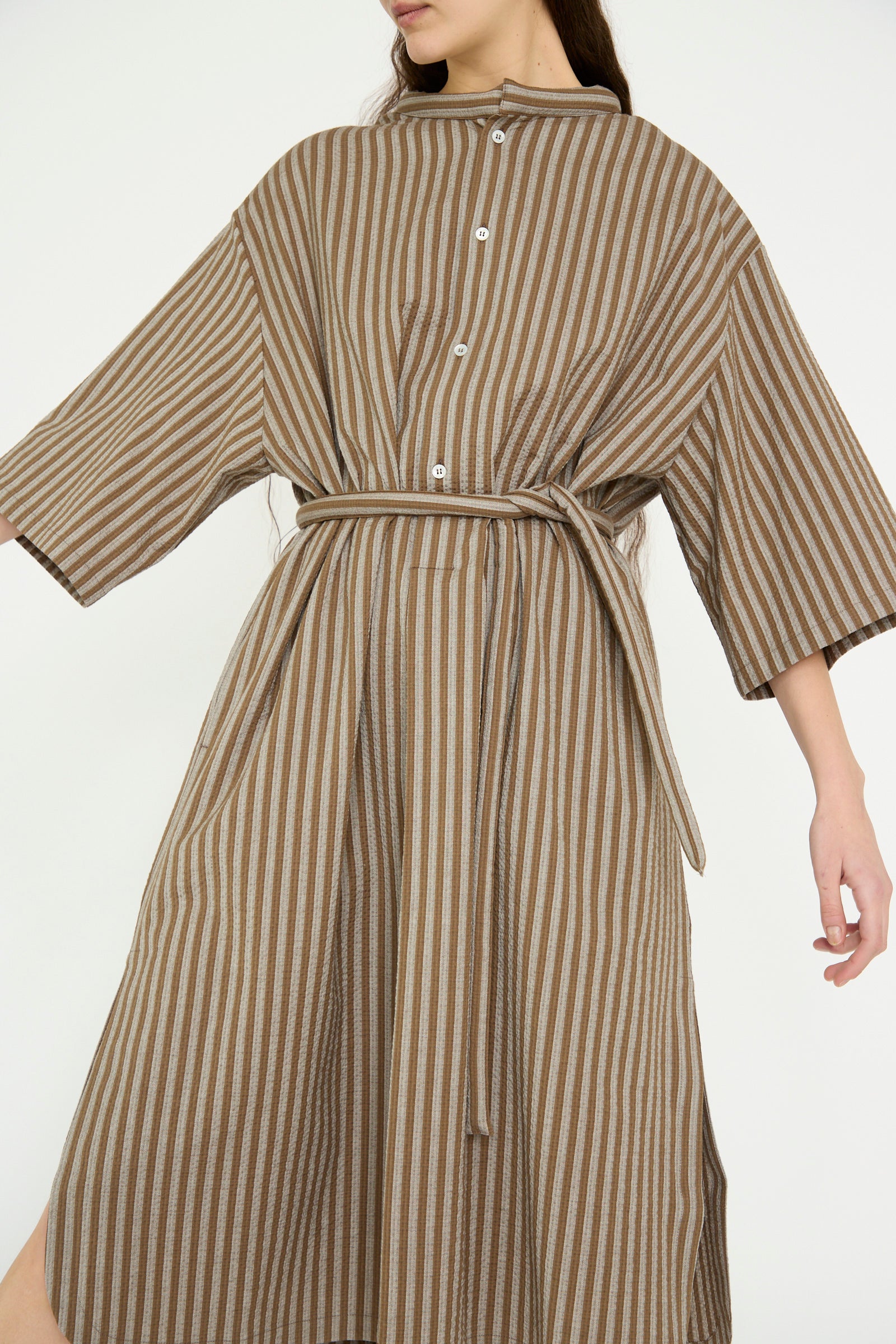 Wearing Cristaseya's brown-striped Caftan with Padded Collar, featuring long sleeves and a waist tie, a model stands  against a plain backdrop.