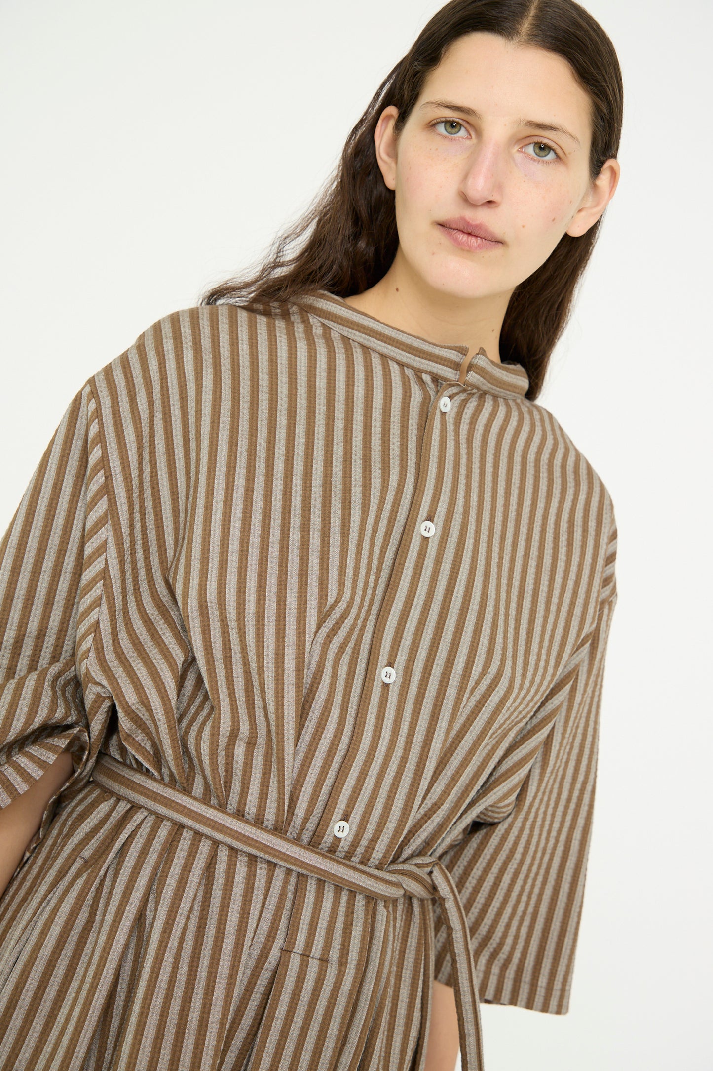 A model with long hair wears the Cristaseya Caftan with Padded Collar in brown stripes, featuring an oversized fit as they stand against a plain background.