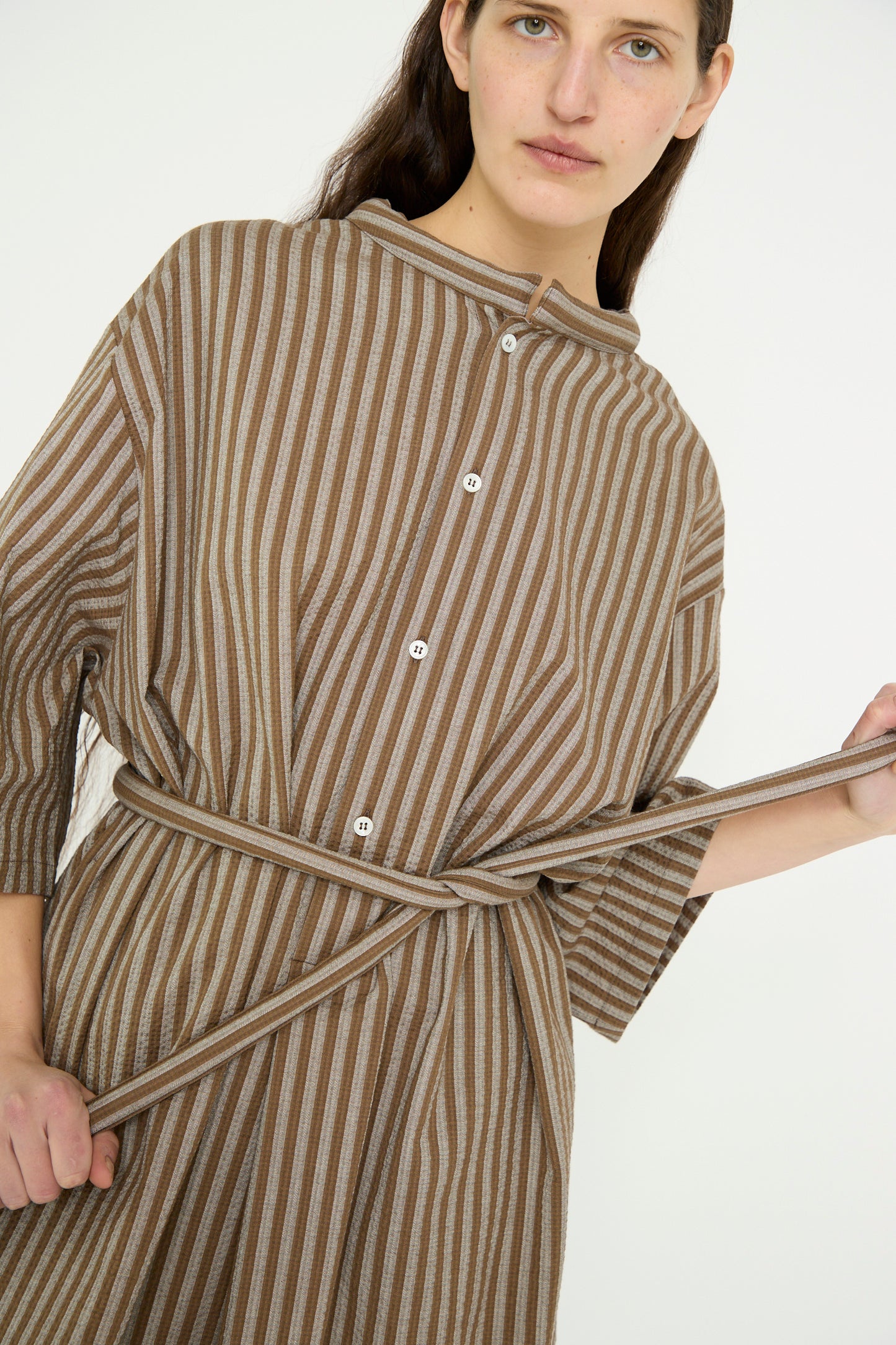 The model is wearing the Cristaseya Caftan with Padded Collar in Brown Stripes, and holding the self-tie belt against a plain background. 