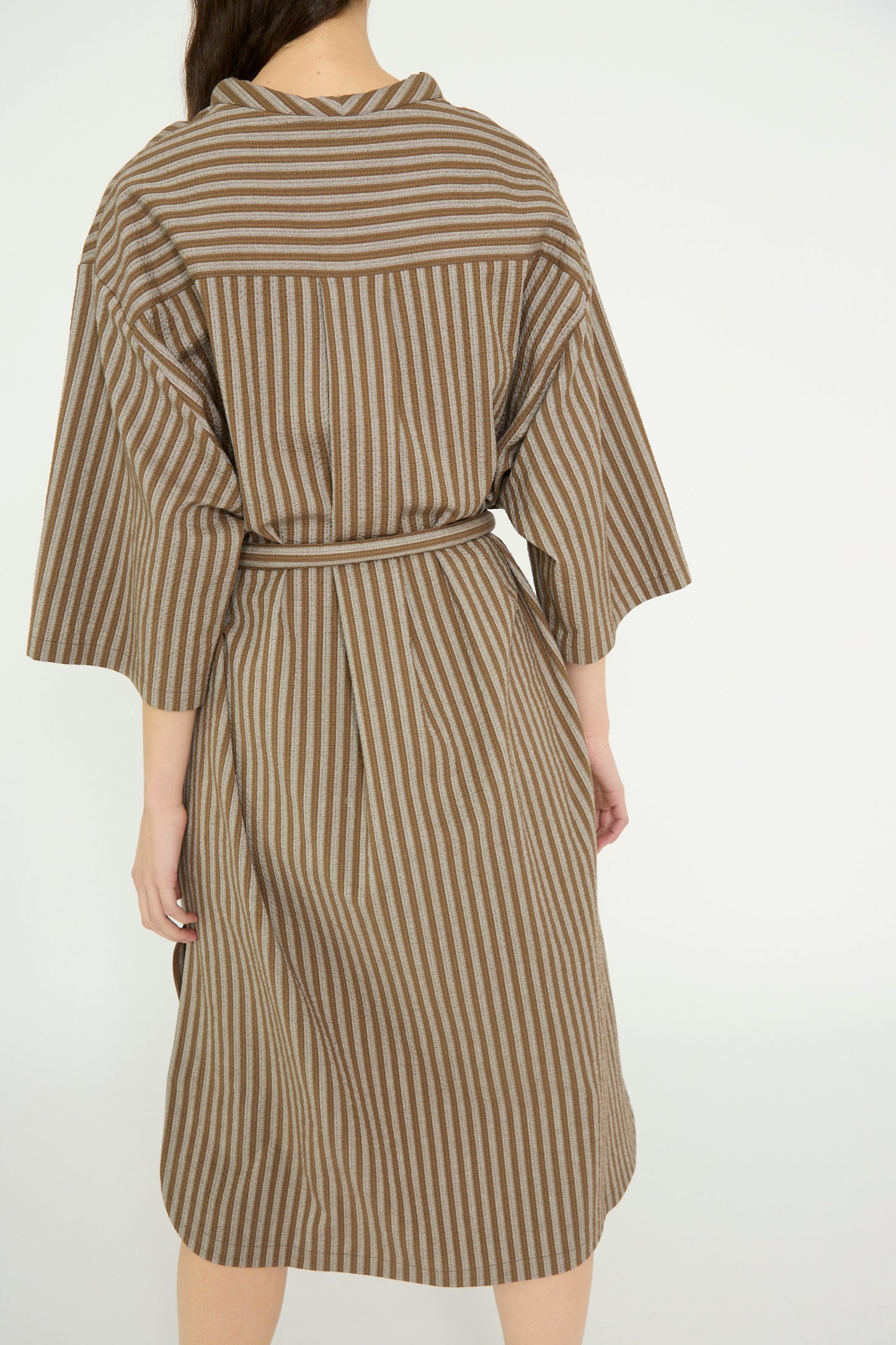 Rear view of a model in an oversized Cristaseya Caftan with Padded Collar, featuring Italian brown and white striped cotton, wide sleeves, and a tied belt, set against a plain background.