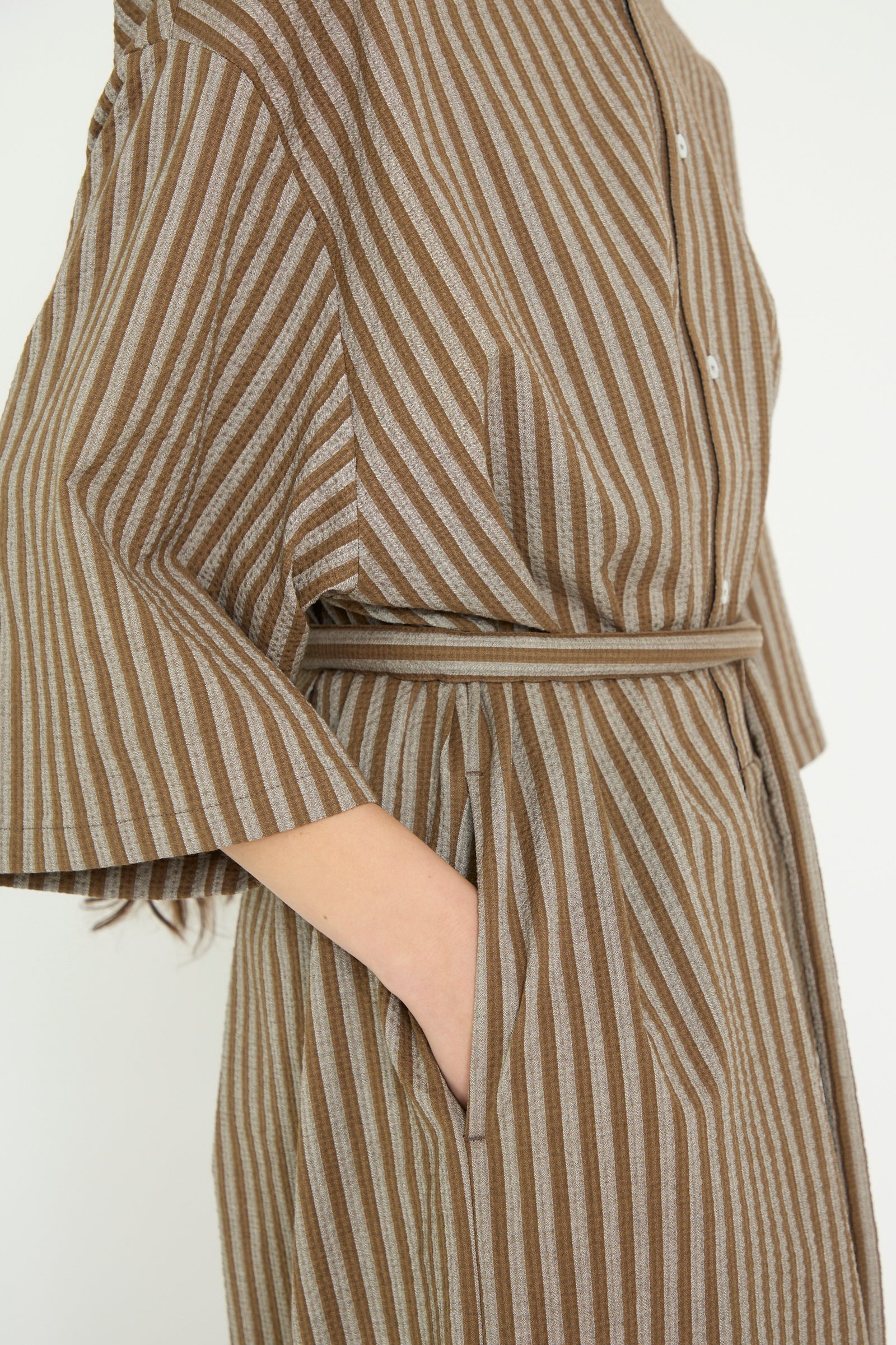 A close-up of a person wearing the Cristaseya Caftan with Padded Collar in Brown Stripes, featuring a belt and short sleeves, with one hand in a side pocket.