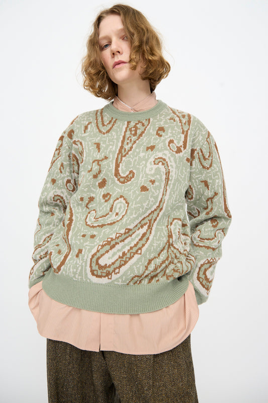 A person with short wavy hair wears the Cristaseya Cashmere Jacquard Sweater in Sauge layered over a pink shirt and brown pants, standing against a plain white background.