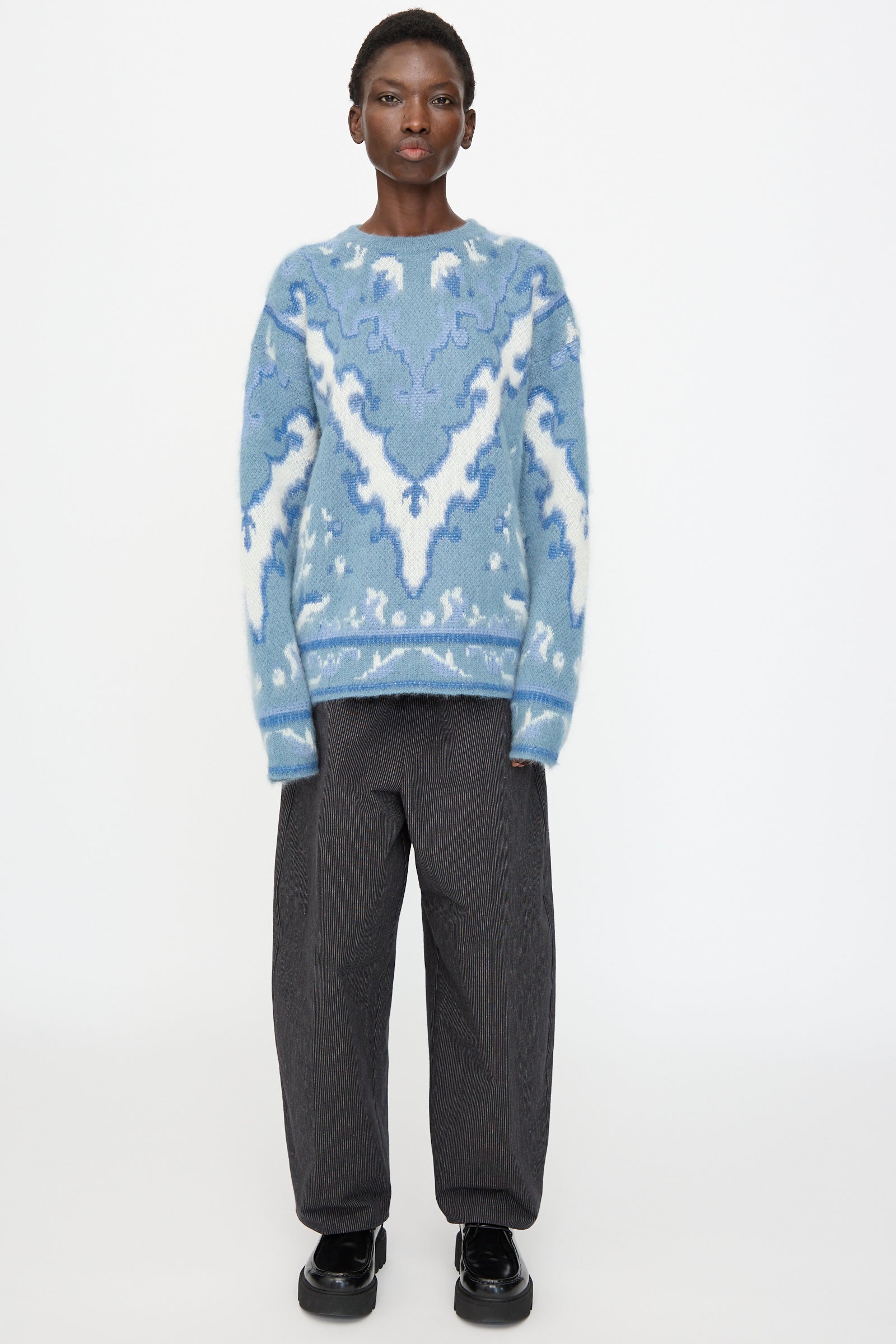 A person stands against a white background wearing Cristaseya’s Cashmere Oversized Carpet Sweater in blue and white, paired with loose dark gray pants and black shoes. The sweater is crafted from a luxurious cashmere wool blend.