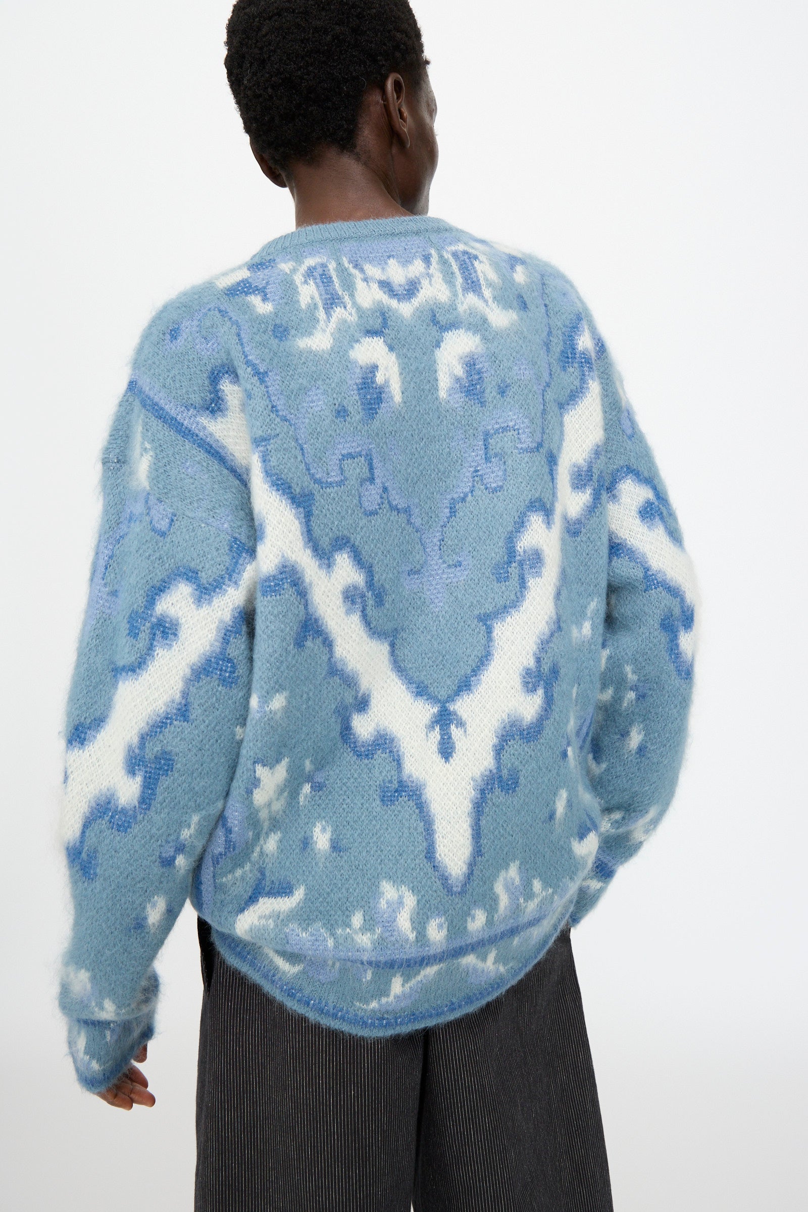 Model wears the Cristaseya Cashmere Oversized Carpet Sweater in Blue and White, styled with dark pinstripe pants, shown from the back against a plain light background.