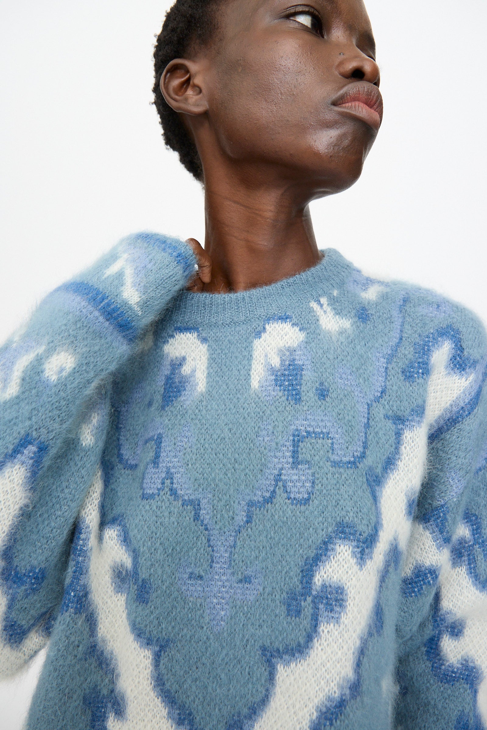 Model wears Cristaseya’s Cashmere Oversized Carpet Sweater in Blue and White, featuring abstract patterns, crafted from a cashmere wool blend, gazing upward against a plain background.