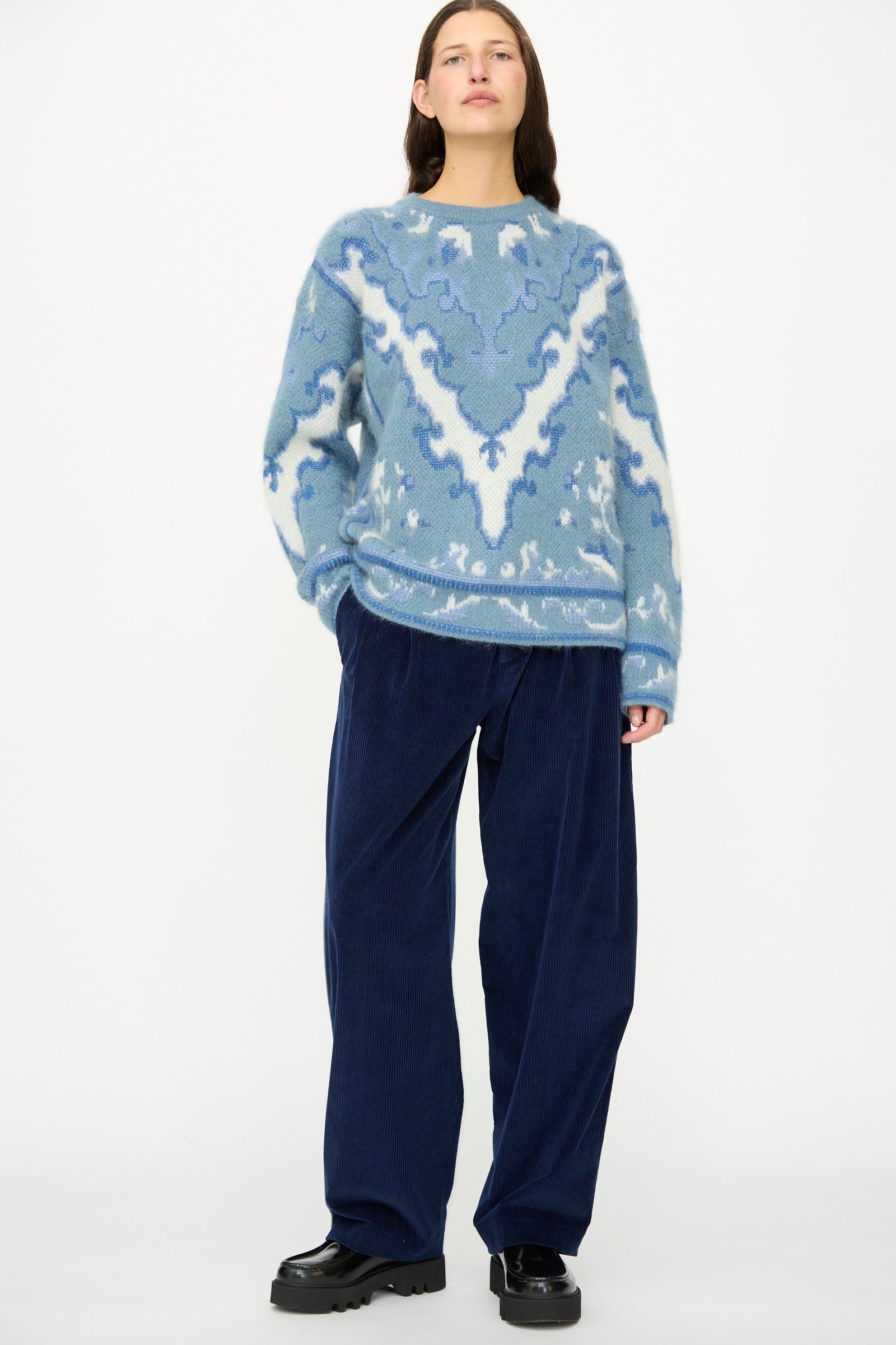 Person standing against a white background, wearing a blue patterned sweater, Cristaseya Corduroy Double Pleated Wide Pants in Indigo, and black shoes.
