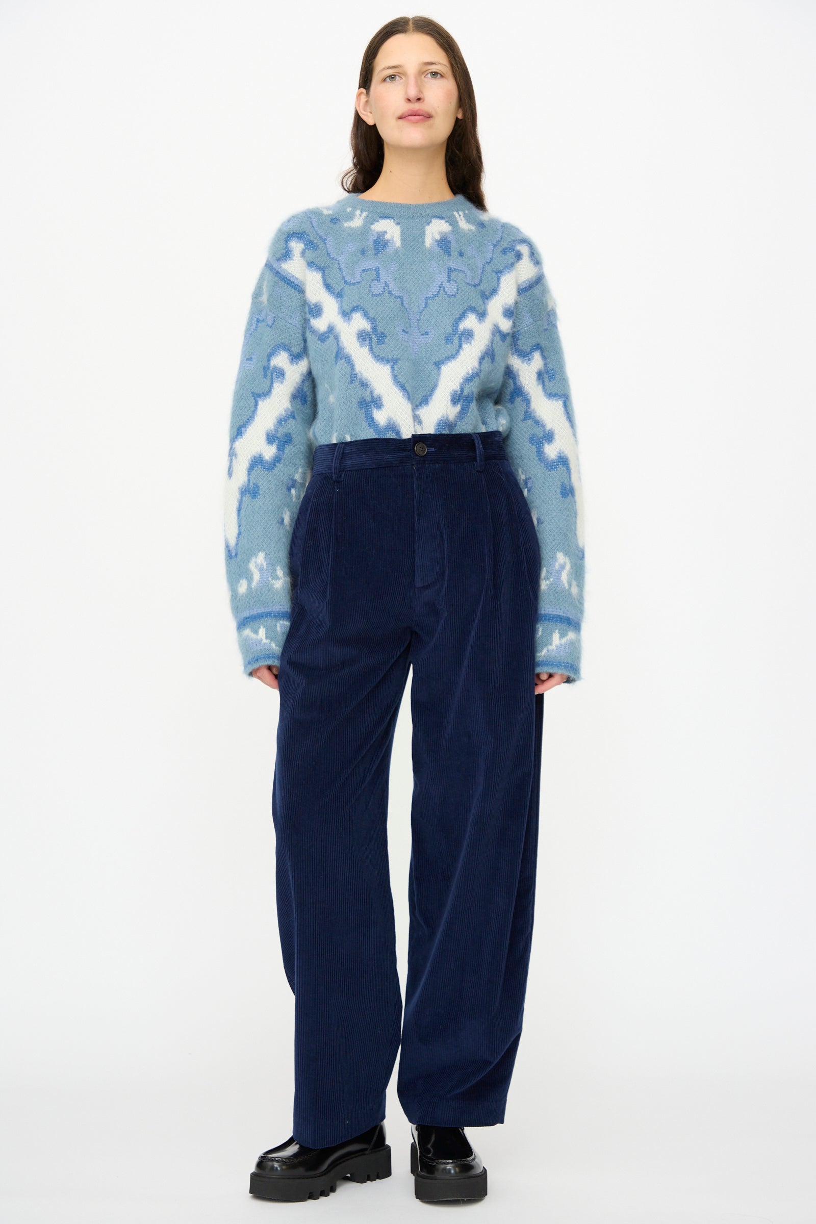 A woman stands against a white background wearing a blue patterned sweater, Cristaseya Corduroy Double Pleated Wide Pants in Indigo, and black shoes.