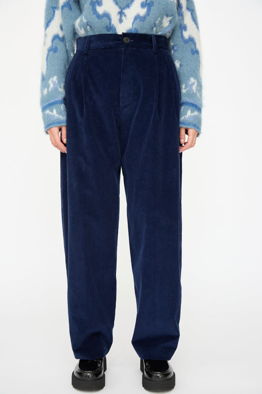 Model wears Cristaseya Corduroy Double Pleated Wide Pants in Indigo, a blue patterned sweater, and black shoes, shown from shoulders to feet against a plain white background.
