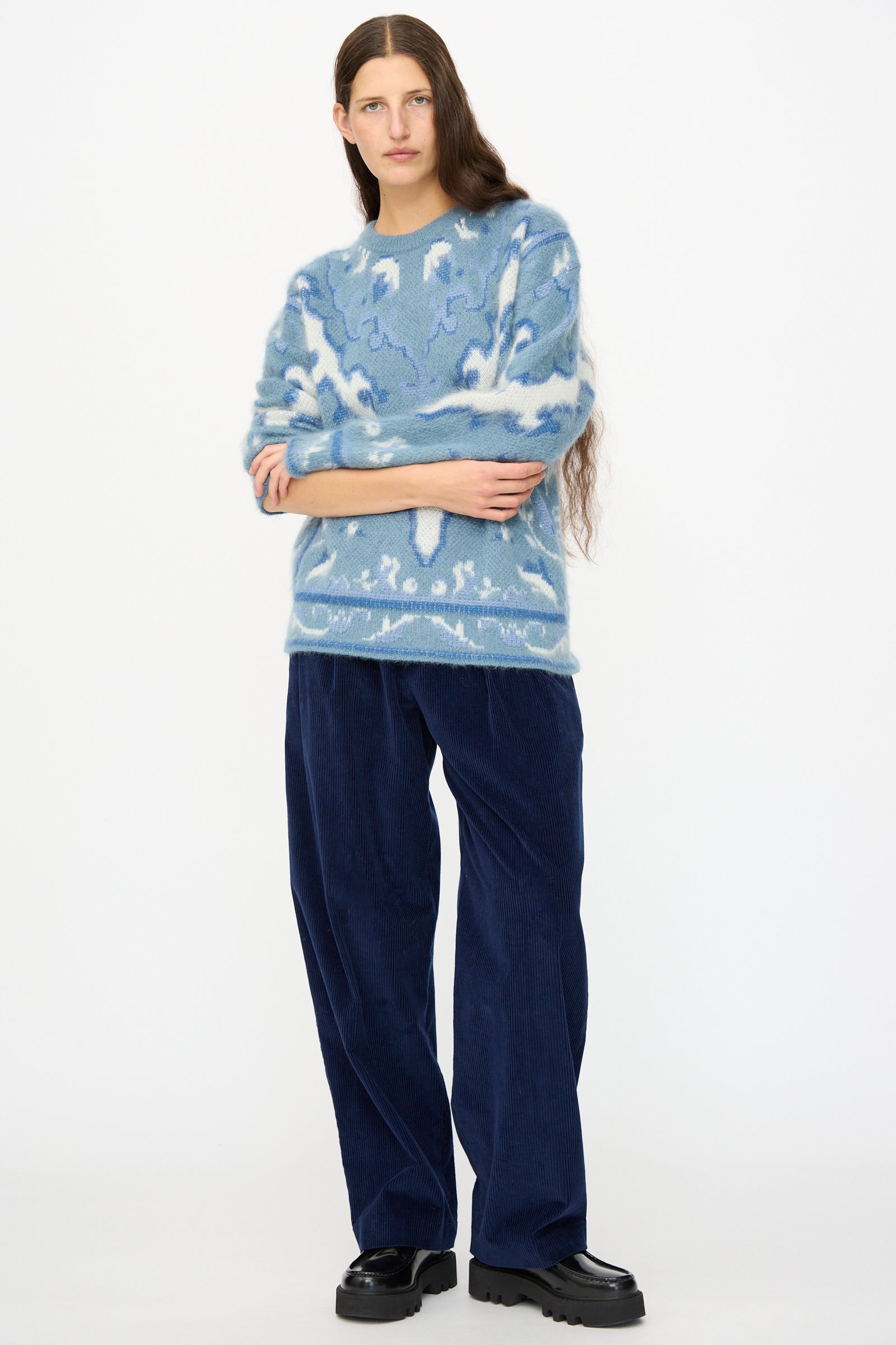 A person with long hair wears a blue patterned sweater, Cristaseya Corduroy Double Pleated Wide Pants in Indigo, and black platform shoes, standing against a plain white background.