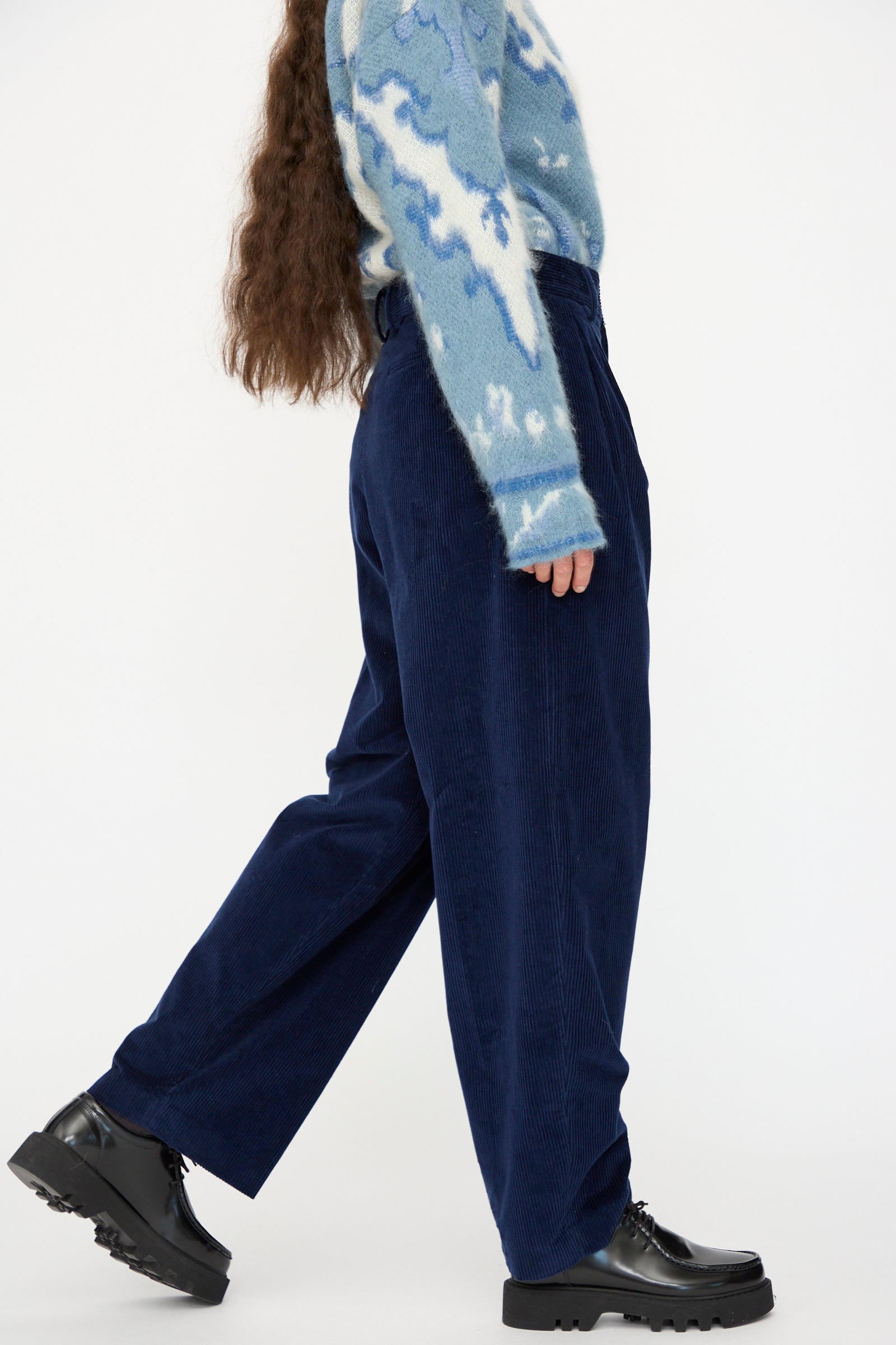 A person walks against a plain white background wearing a blue and white patterned sweater, Cristaseya Corduroy Double Pleated Wide Pants in Indigo, and black platform shoes.
