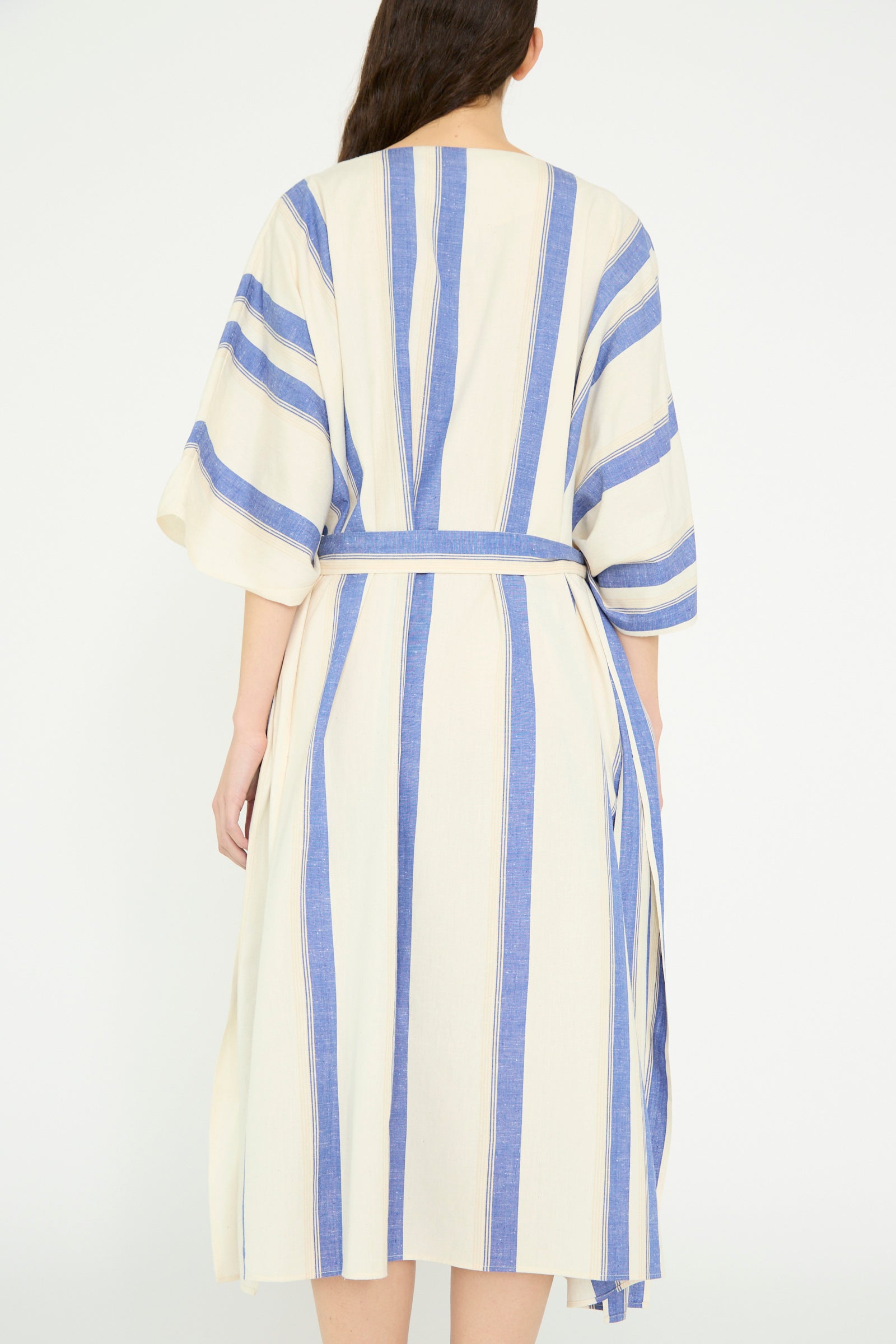 Rear view of a person wearing Cristaseya's Cotton Maxi Caftan featuring vertical blue stripes on white, with wide sleeves and a cinched waist tie against a white backdrop. 