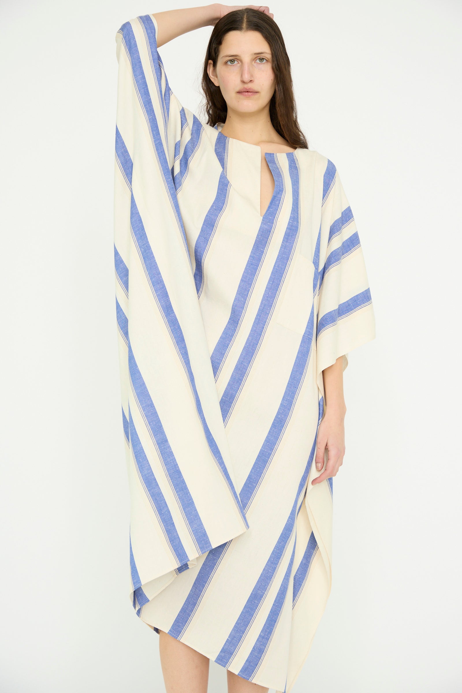 A model poses in a Cristaseya Cotton Maxi Caftan featuring diagonal blue and white stripes, one hand resting on their head against a plain backdrop.