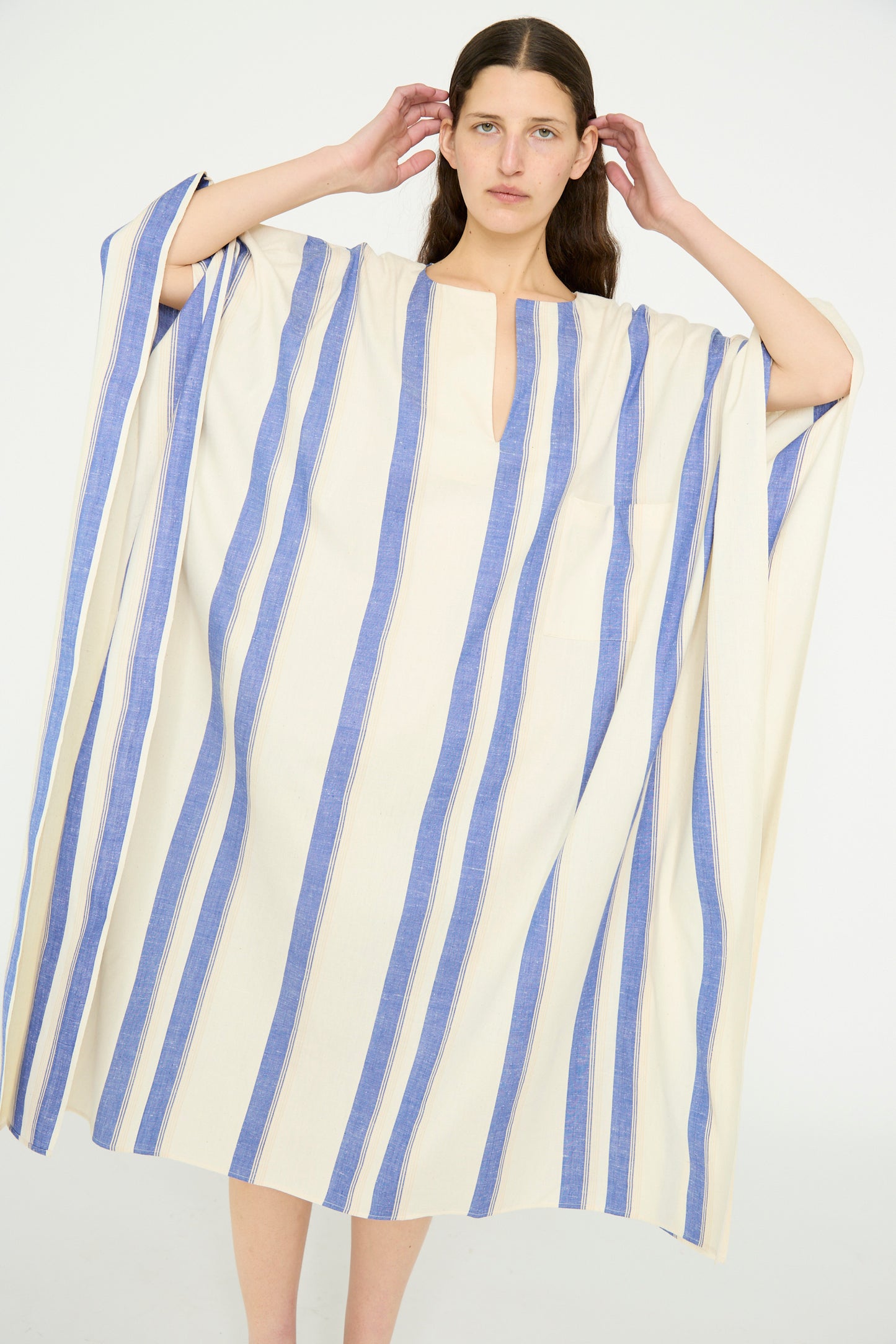 A model stands adjusting their hair against a plain background, wearing the Cristaseya Cotton Maxi Caftan in White with Blue Stripes.