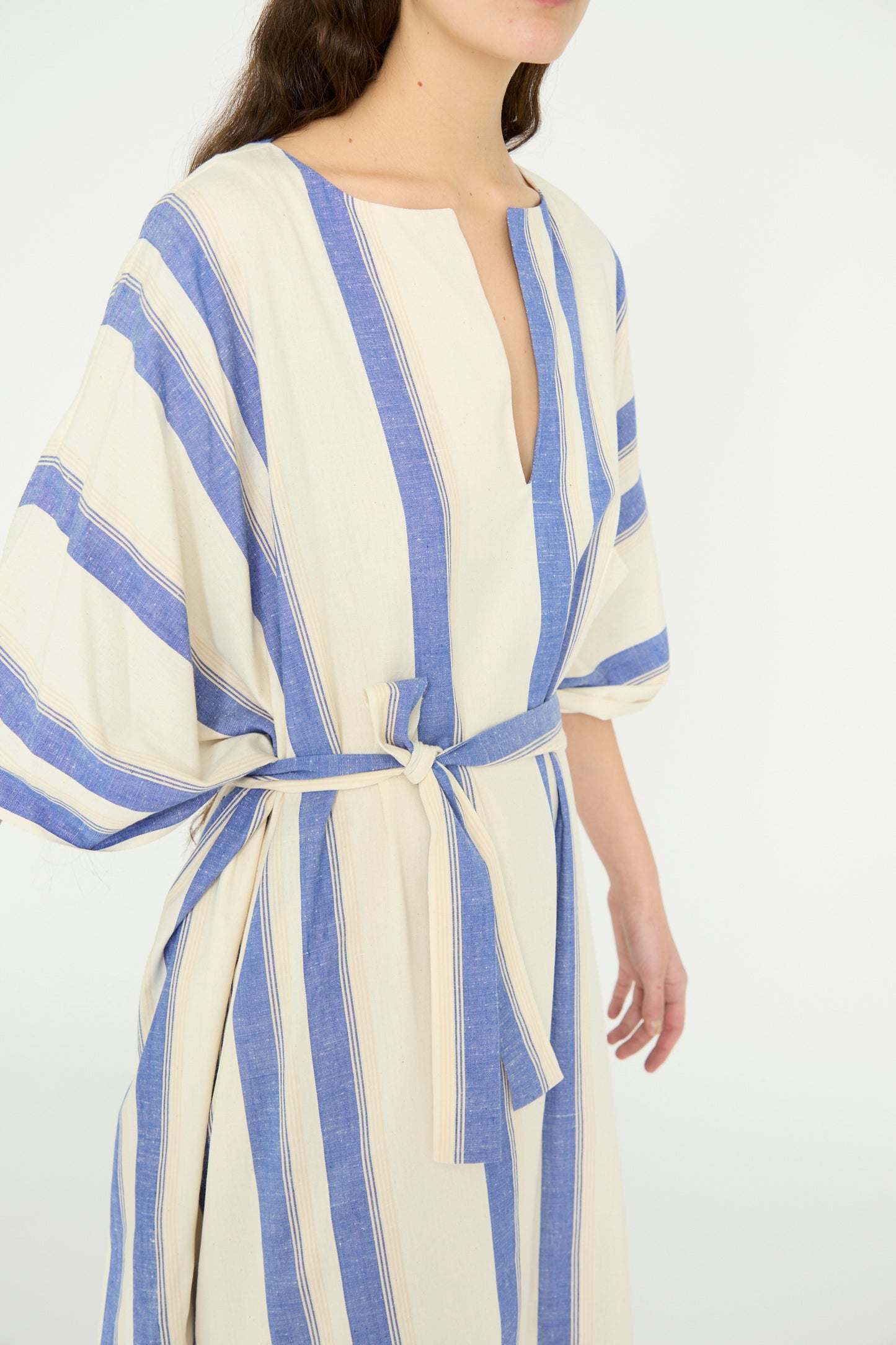 A model wearing the Cristaseya Cotton Maxi Caftan in White with Blue Stripes, featuring wide sleeves, a waist tie, and an oversized fit against a plain background, seen from below the chin. 