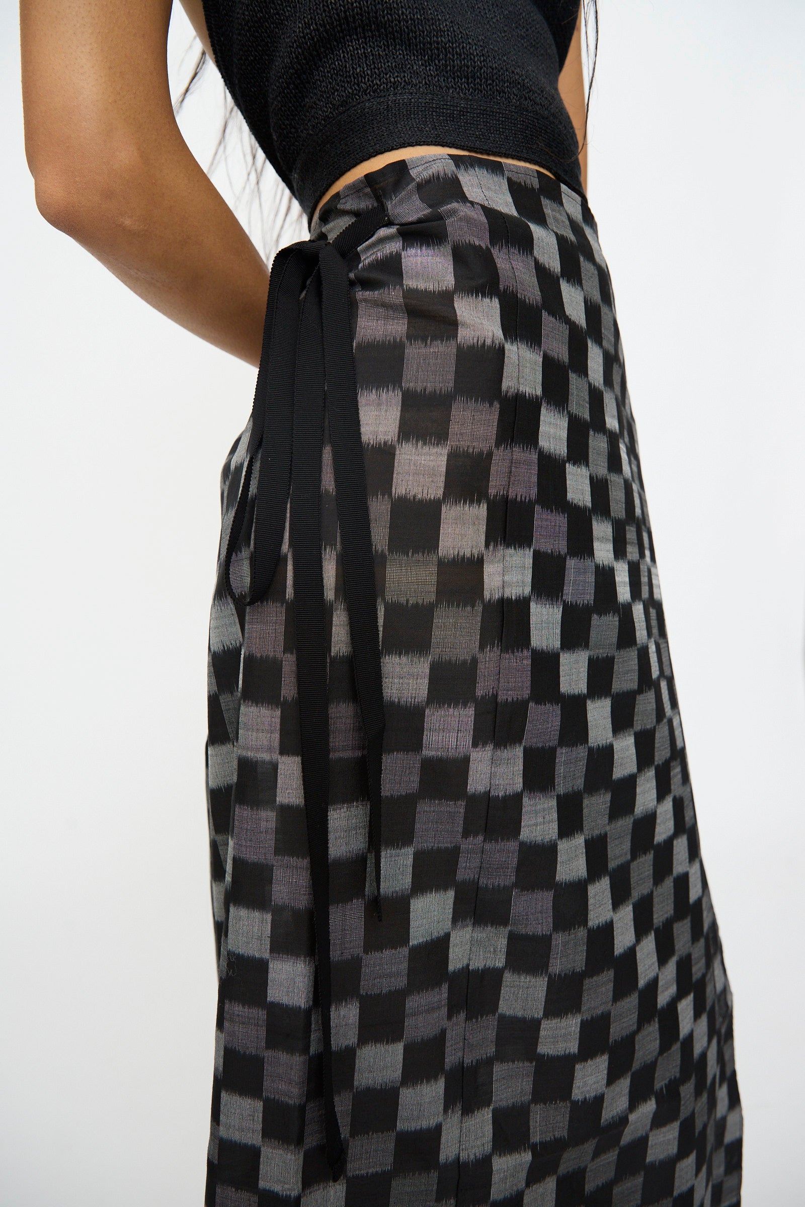 A person wearing Cristaseya's Cotton Silk Ikat Pareo Skirt in Black Grey Checks, shown from the waist down, paired with a black sleeveless top.