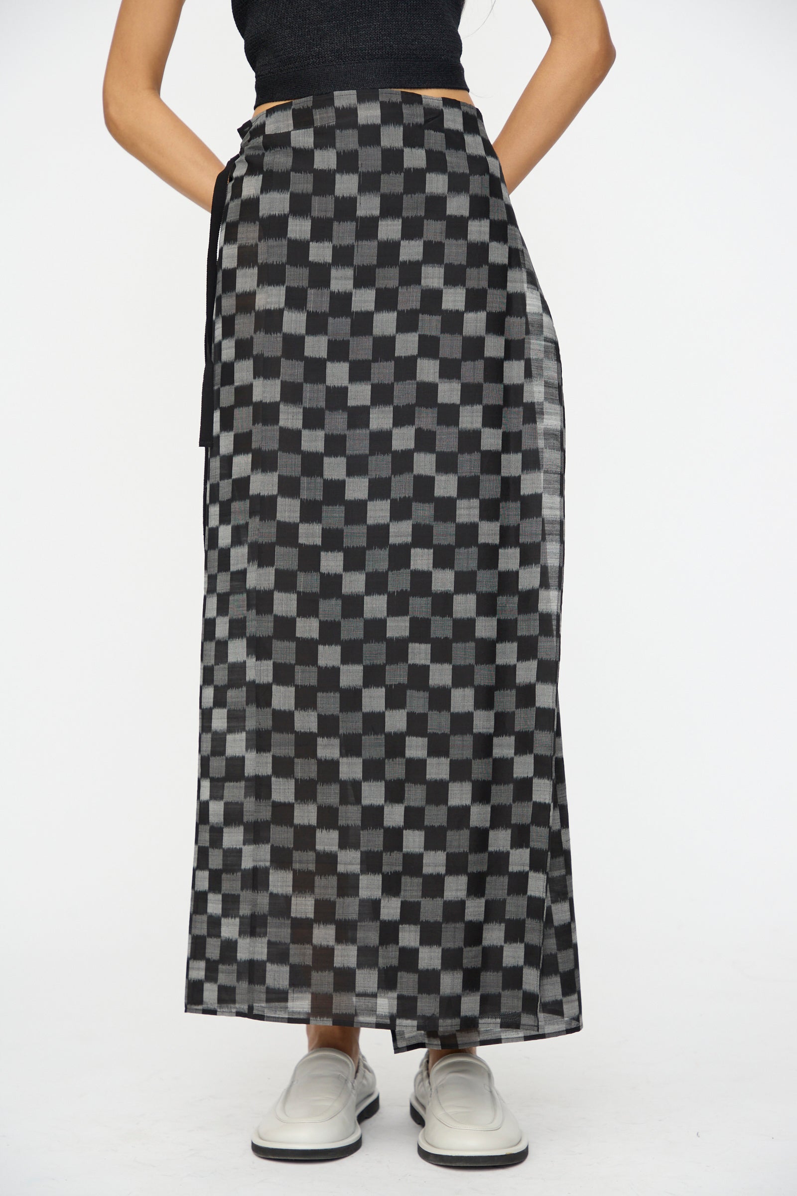 A person models the Cristaseya Cotton Silk Ikat Pareo Skirt in Black Grey Checks, styled with a black top and white slip-on shoes, against a plain white background.