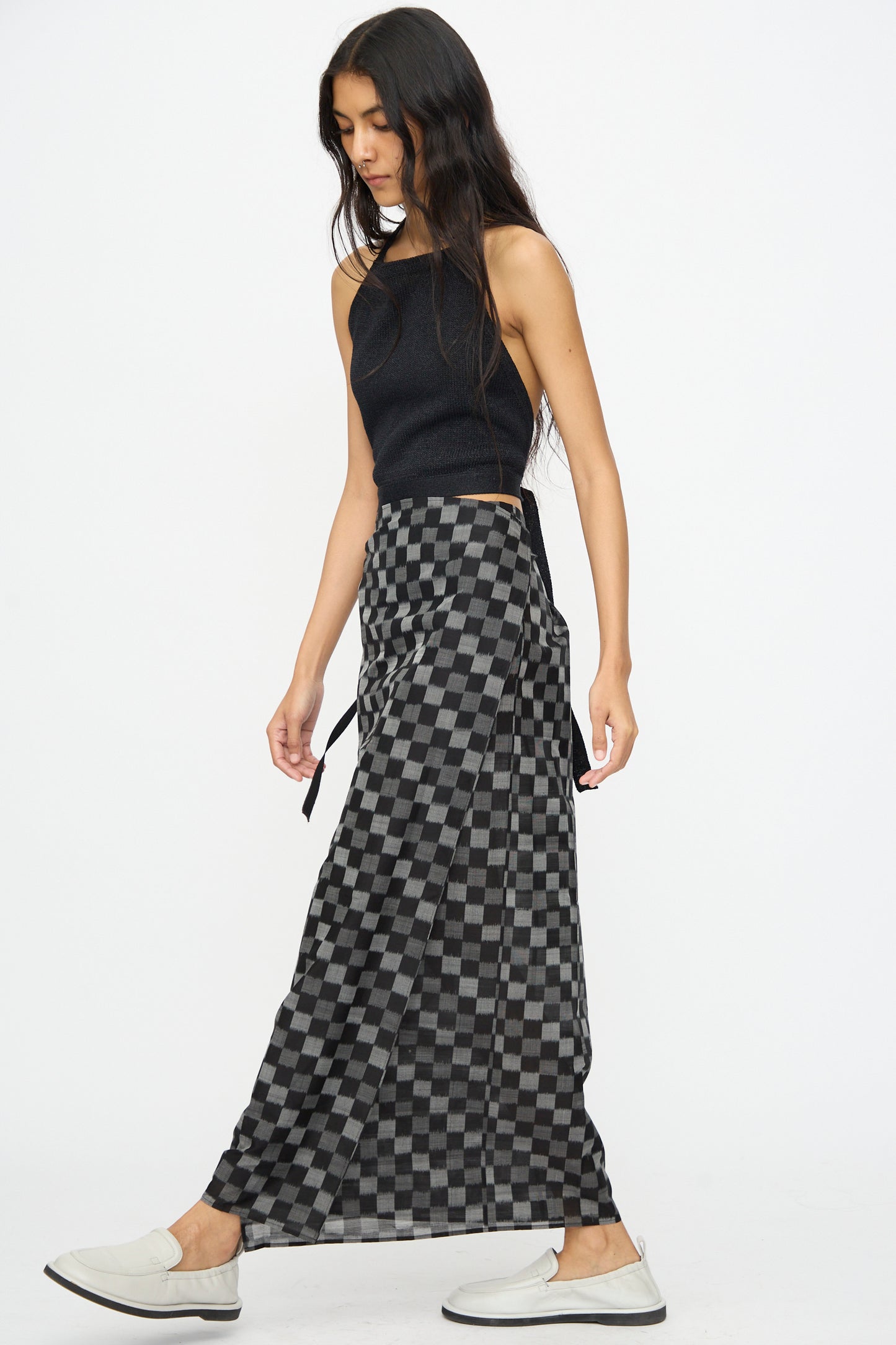 A woman walks sideways in a Cristaseya Cotton Silk Ikat Pareo Skirt in Black Grey Checks, paired with a black sleeveless top and white slip-on shoes, set against a plain white background.