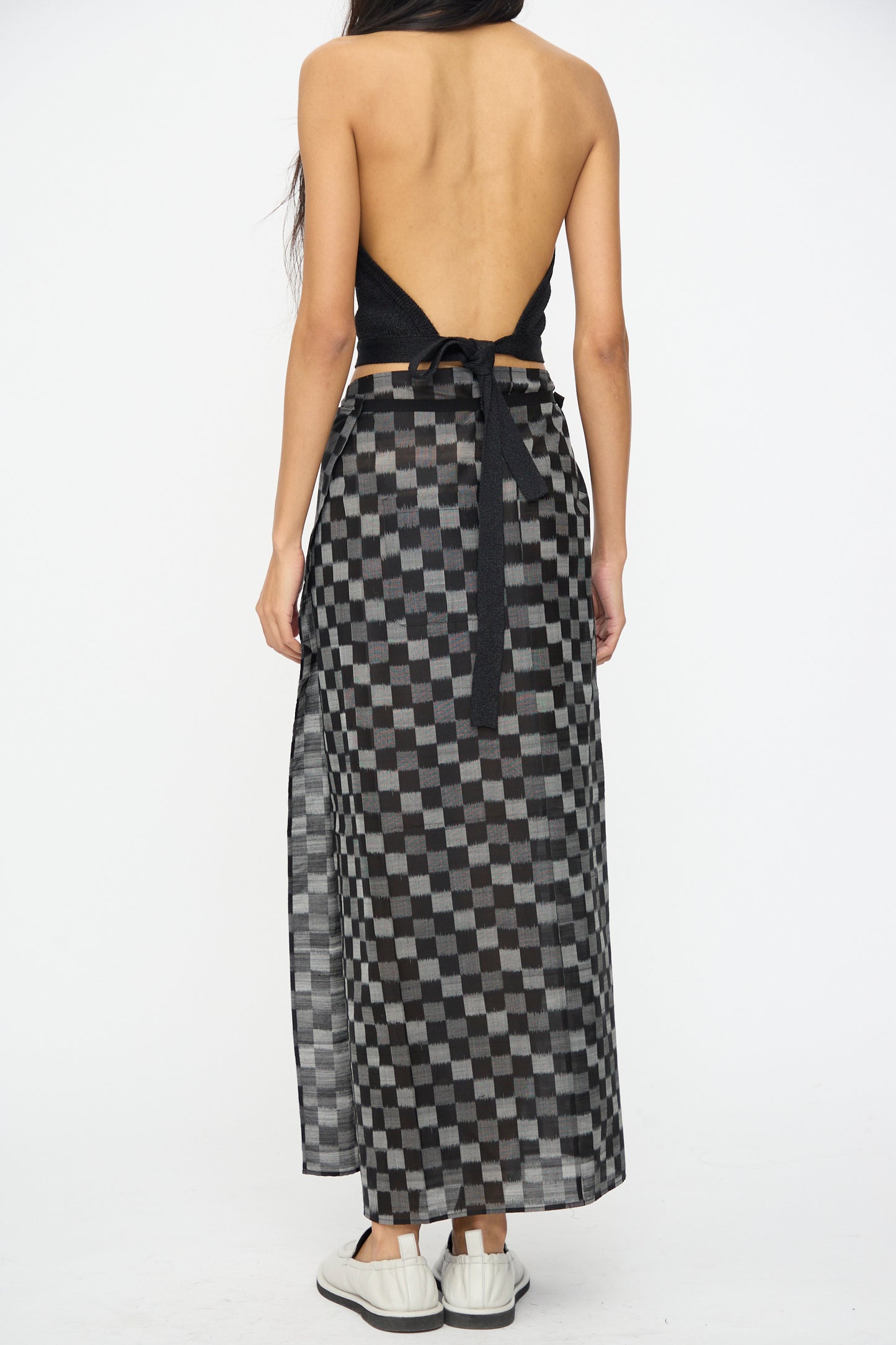 A woman faces away, wearing a black halter backless top with the Cristaseya Cotton Silk Ikat Pareo Skirt in Black Grey Checks, paired with white sneakers.