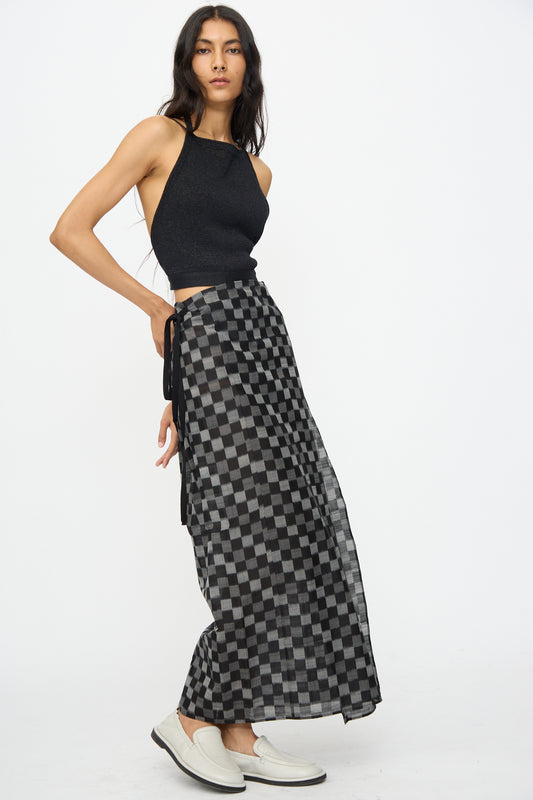 A woman wearing a sleeveless black top, Cristaseya’s Cotton Silk Ikat Pareo Skirt in Black Grey Checks, and white loafers stands against a plain white background.