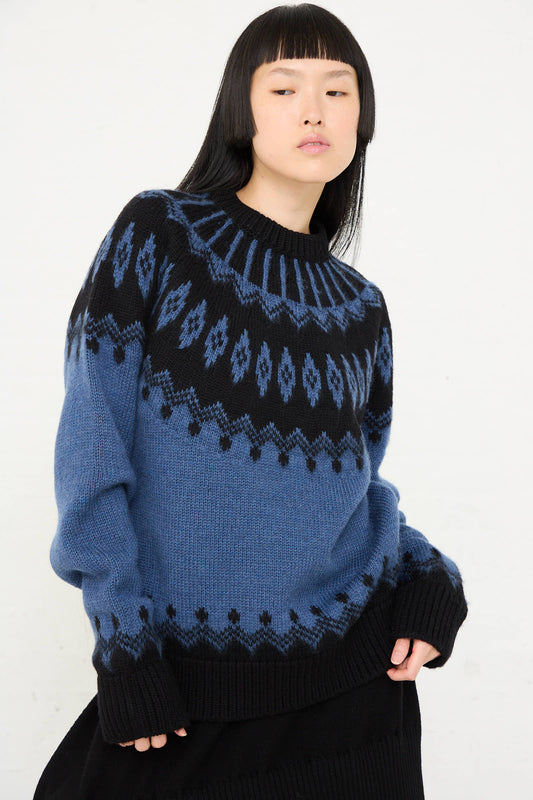 A person with long black hair is wearing the Cristaseya Extra Fine Merino Oversized Mountain Sweater in Black, which features a blue and black geometric design. They are standing against a plain white background.