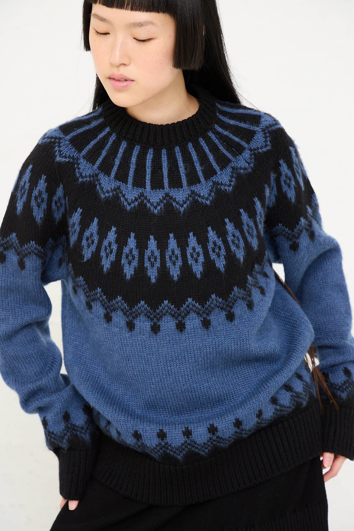 Person wearing the Cristaseya Extra Fine Merino Oversized Mountain Sweater in Black, featuring a blue and black mountain motif, hands in pockets, looking down.