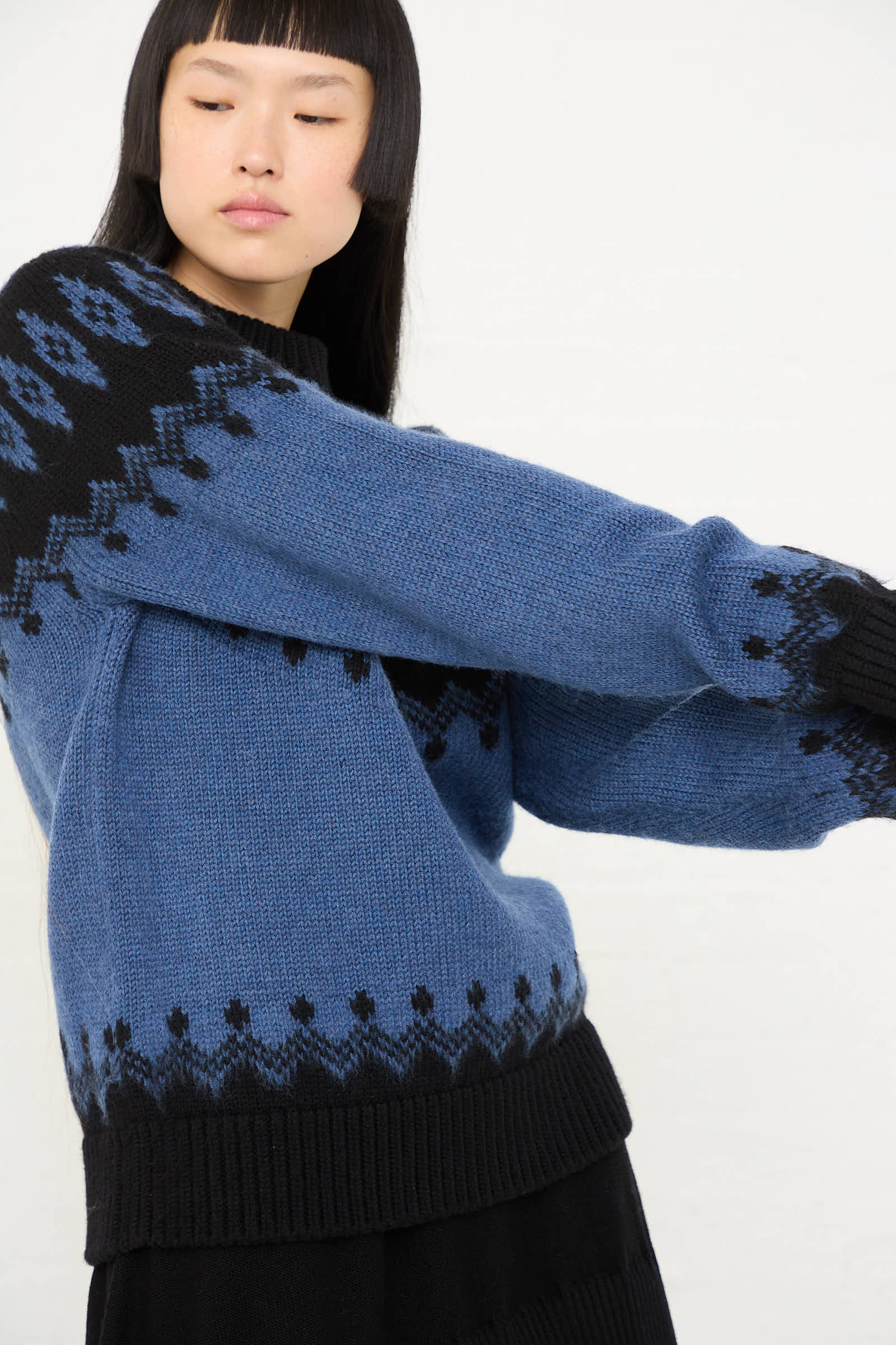 A person wearing Cristaseya's Extra Fine Merino Oversized Mountain Sweater in Black, featuring a blue and black mountain motif and geometric pattern, looking to the side with arms extended.