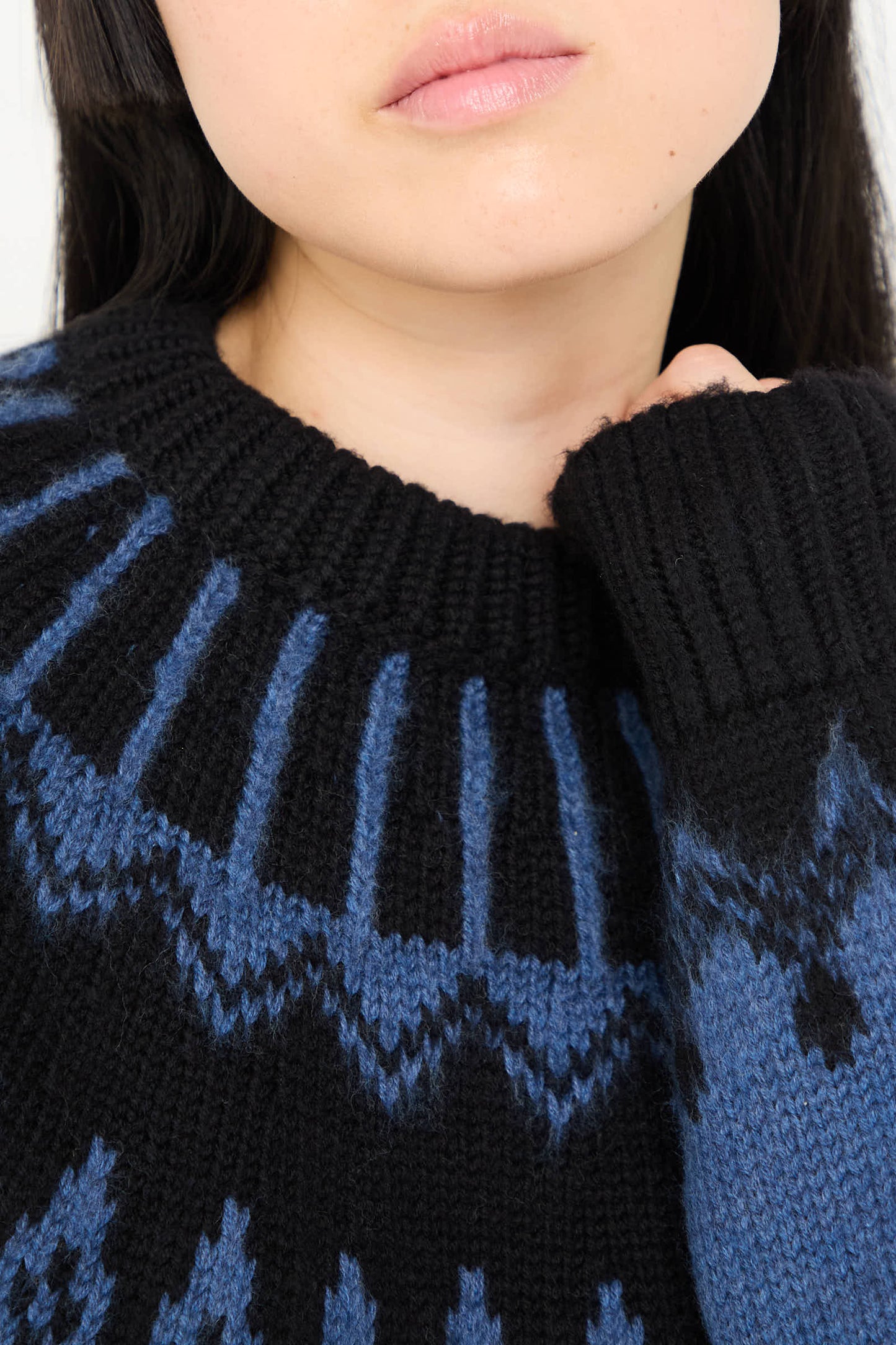 Close-up of a person wearing an Extra Fine Merino Oversized Mountain Sweater in Black by Cristaseya, featuring a black and blue pattern. Only their lower face and part of their black hair are visible, along with their hand tugging at the collar.