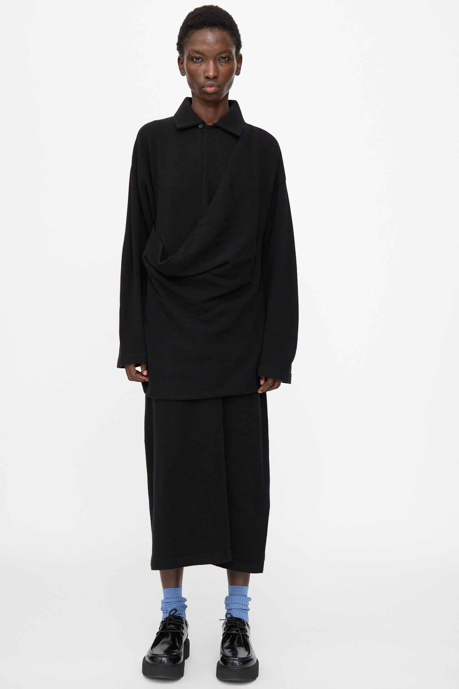 A person stands against a white background wearing the Cristaseya Flannel Polo Dress in Black, paired with blue socks and black shoes.