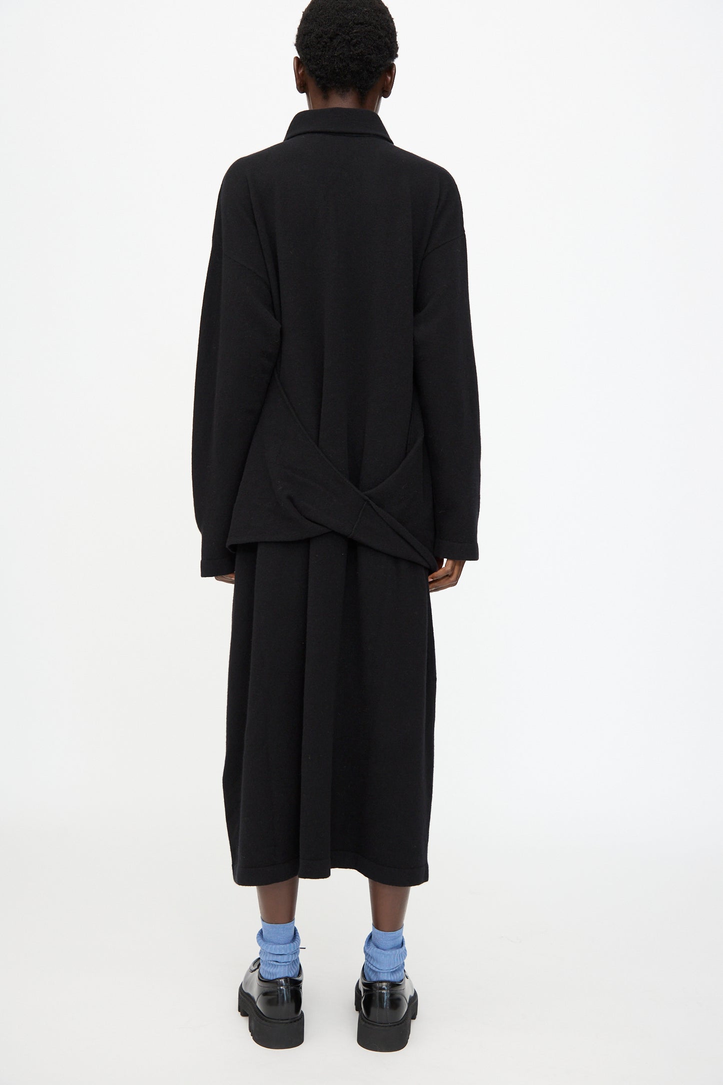 A person is shown from the back against a white background, wearing the Cristaseya Flannel Polo Dress in Black styled as a long coat, with a black skirt, blue socks, and black platform shoes.