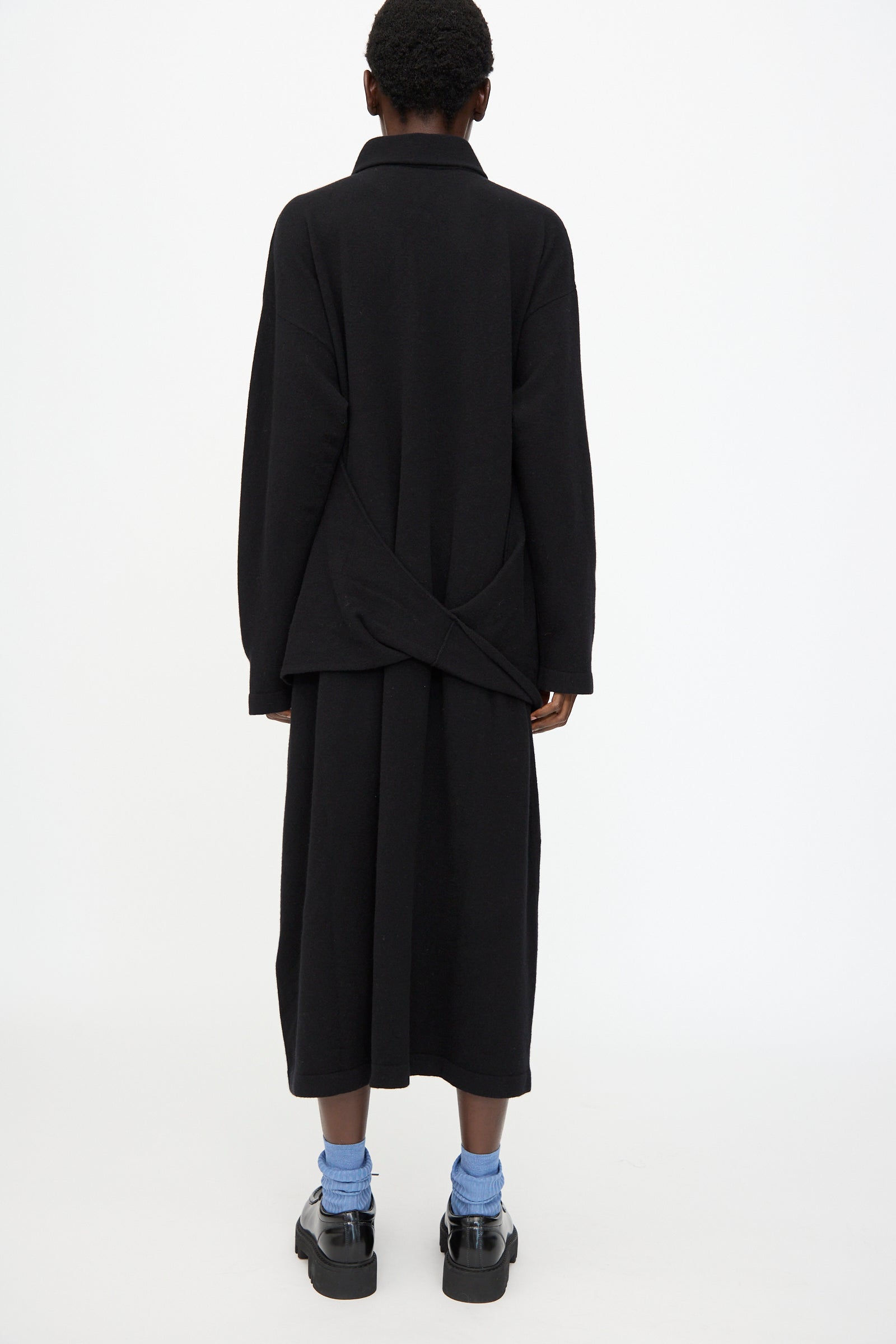 A person is shown from the back against a white background, wearing the Cristaseya Flannel Polo Dress in Black styled as a long coat, with a black skirt, blue socks, and black platform shoes.