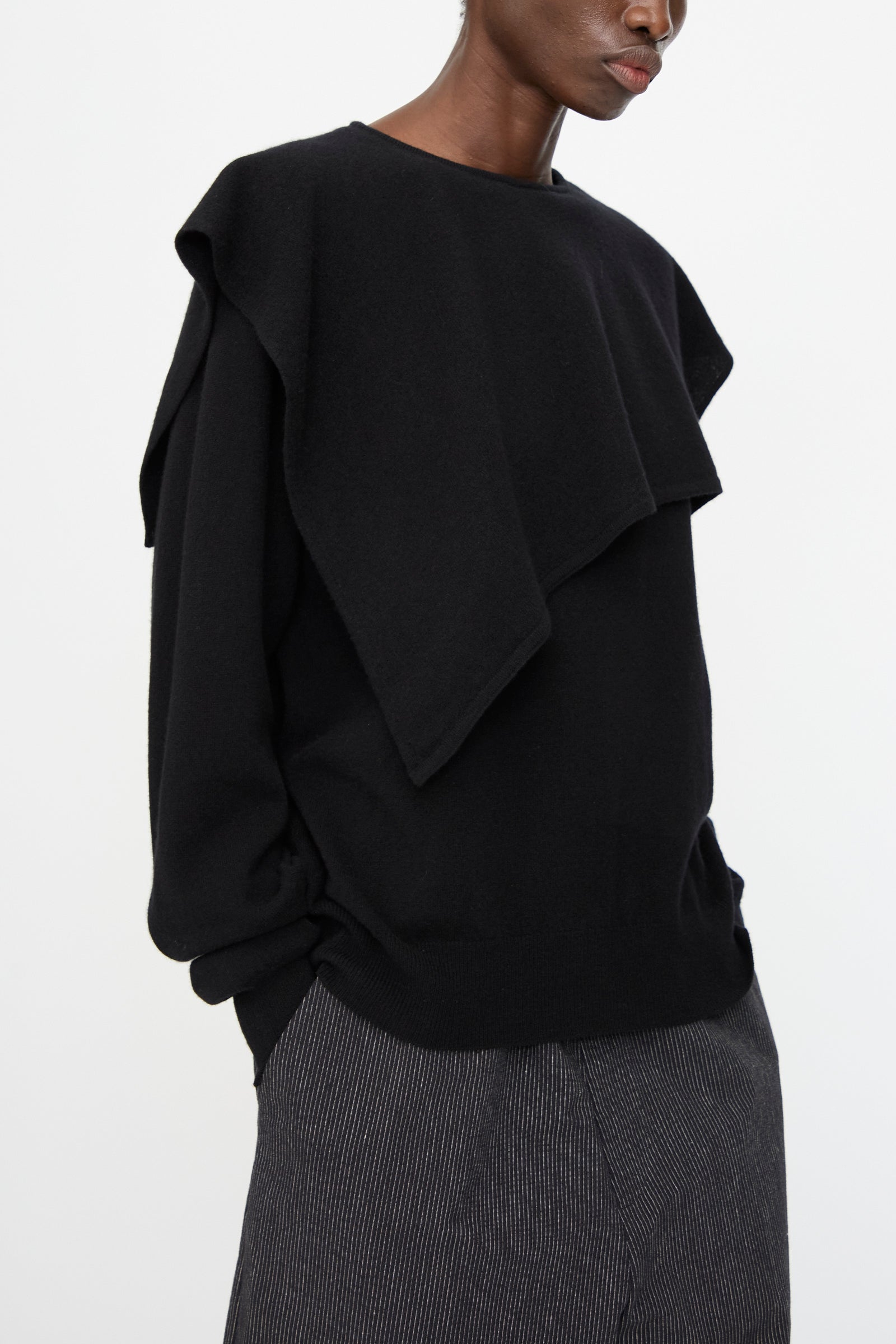 A person wears the Cristaseya Flannel Sailor Collar Sweater in Black, featuring layered fabric over the shoulders, paired with gray pinstriped pants, and stands against a plain background.