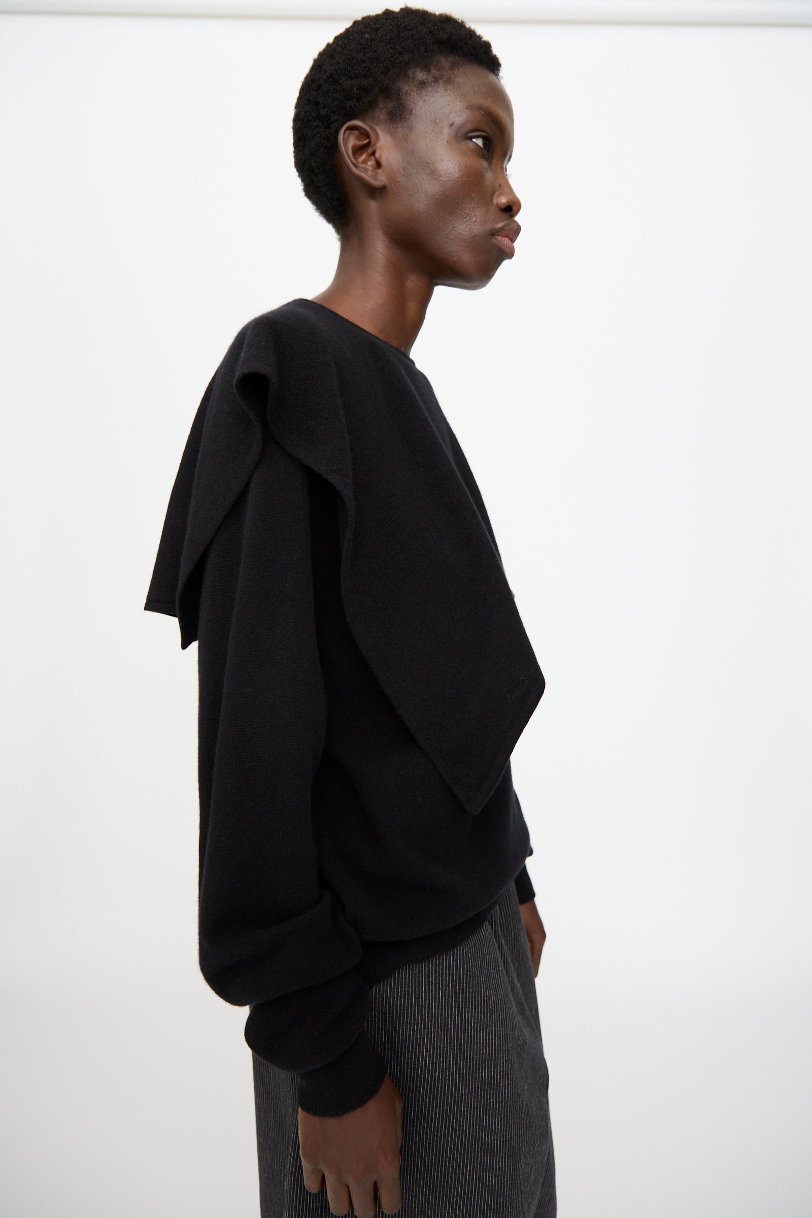 A person in profile stands against a plain background, wearing Cristaseya’s Flannel Sailor Collar Sweater in Black with extra black fabric over one shoulder, paired with striped pants.