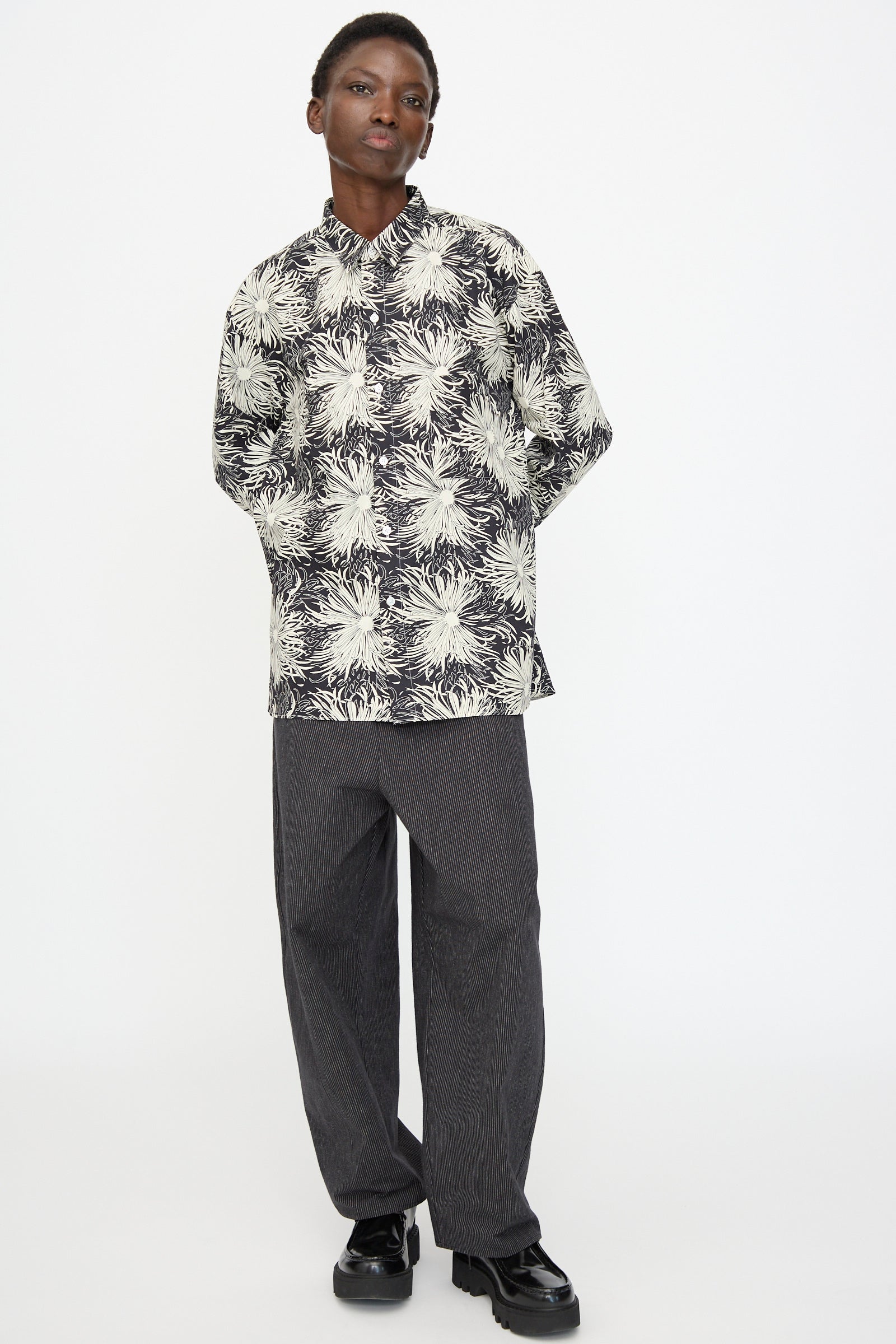 A person stands against a plain white background wearing the Cristaseya Handmade Classic Collar Shirt in Black with White Flowers, paired with loose dark gray pants and black shoes.