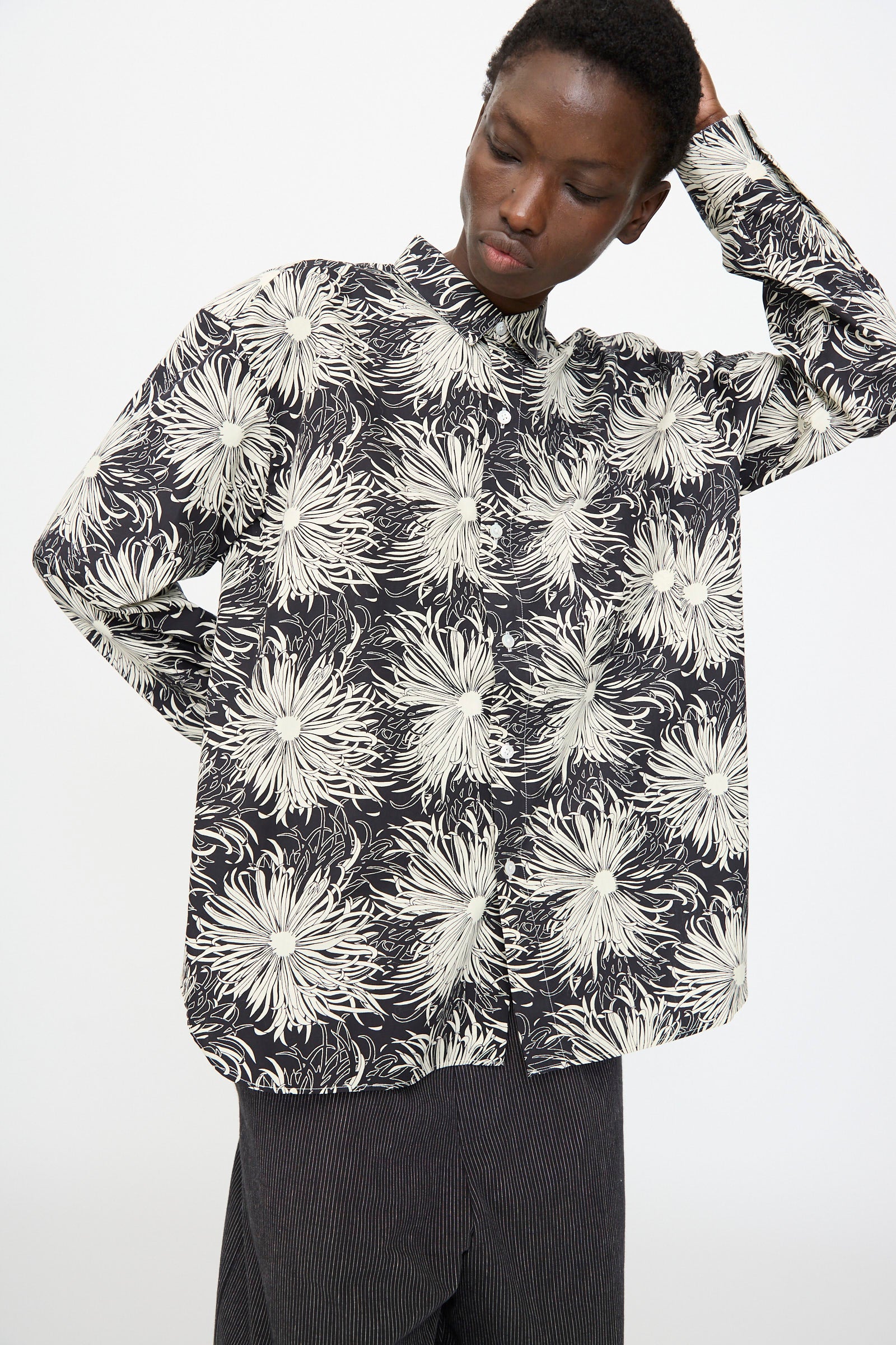 A person wears the Cristaseya Handmade Classic Collar Shirt in Black with White Flowers, paired with dark pants, standing against a plain background with one hand behind their head.