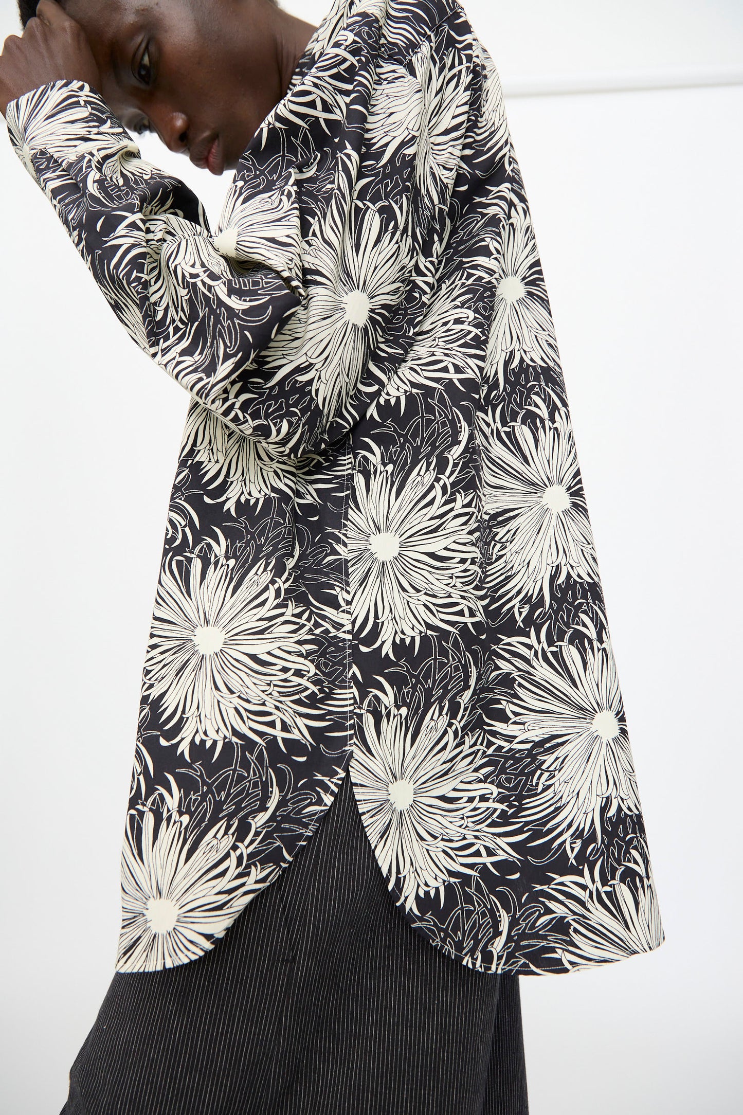 A person models the Cristaseya Handmade Classic Collar Shirt in Black with White Flowers, pairing it with pinstriped pants and posing with one arm bent and head tilted down.