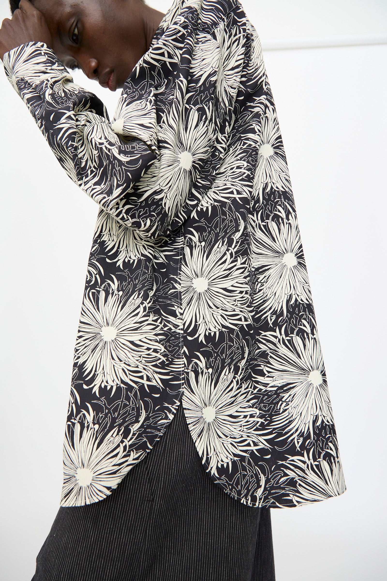 A person models the Cristaseya Handmade Classic Collar Shirt in Black with White Flowers, pairing it with pinstriped pants and posing with one arm bent and head tilted down.