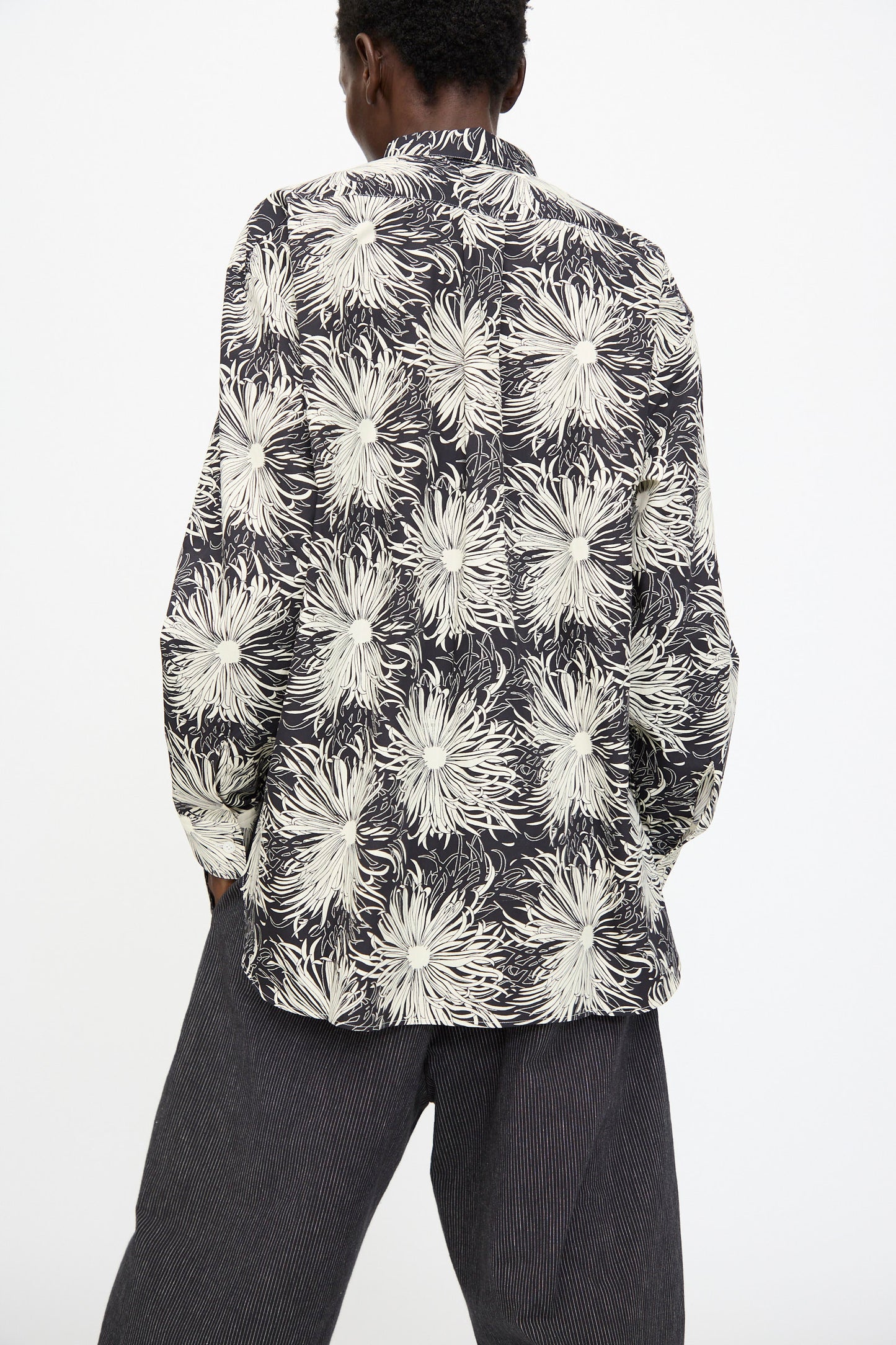 A person stands facing away, wearing the Cristaseya Handmade Classic Collar Shirt in Black with White Flowers and dark pinstriped pants against a plain background.