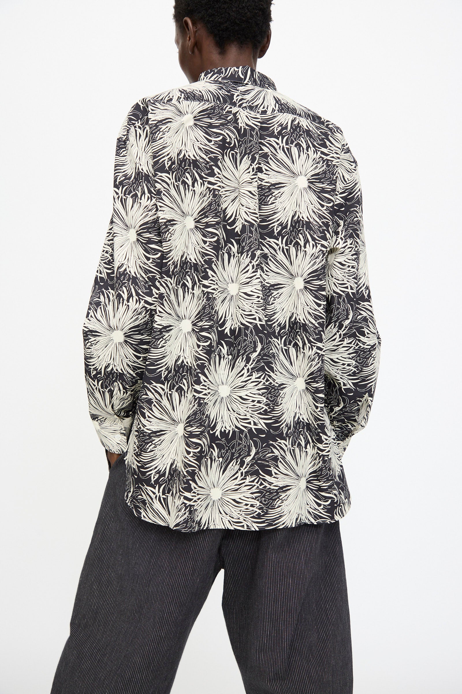 A person stands facing away, wearing the Cristaseya Handmade Classic Collar Shirt in Black with White Flowers and dark pinstriped pants against a plain background.
