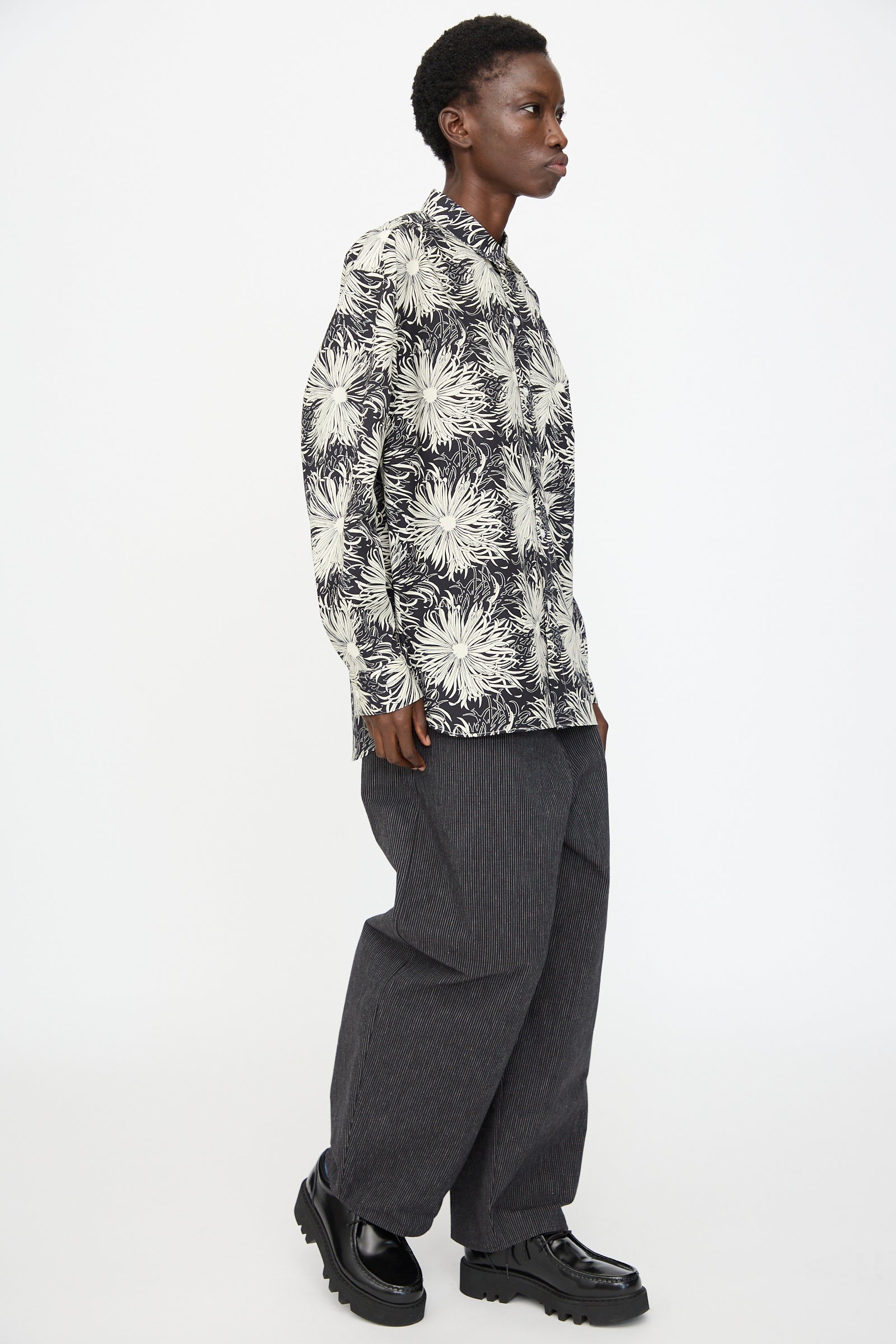 A person in profile wears the Cristaseya Handmade Classic Collar Shirt in Black with White Flowers, wide gray pinstripe pants, and black platform shoes against a plain white background.