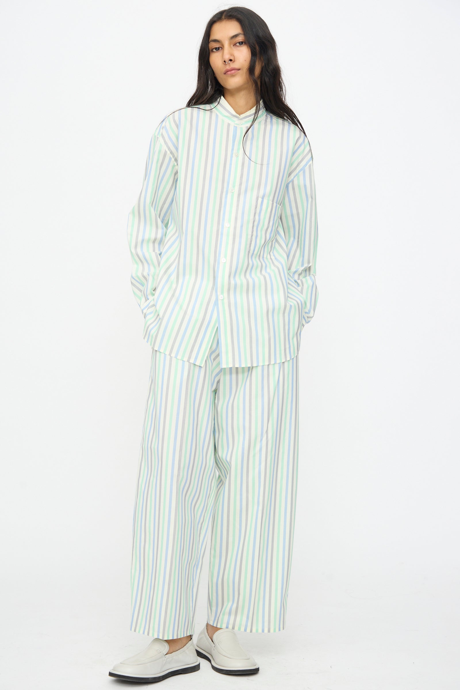 A person stands against a white background wearing the Cristaseya Handmade Fringed Mao Collar Shirt in Striped Pastel Green, styled with matching wide-leg pants and white slip-on shoes.