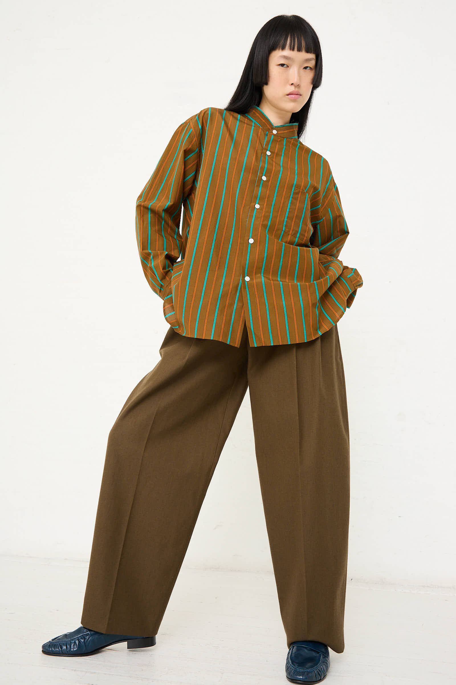 A person stands against a plain background wearing Cristaseya's Handmade Fringed Mao Collar Shirt in Noisette with Blue Stripe and wide-legged pants, with hands in their pockets.