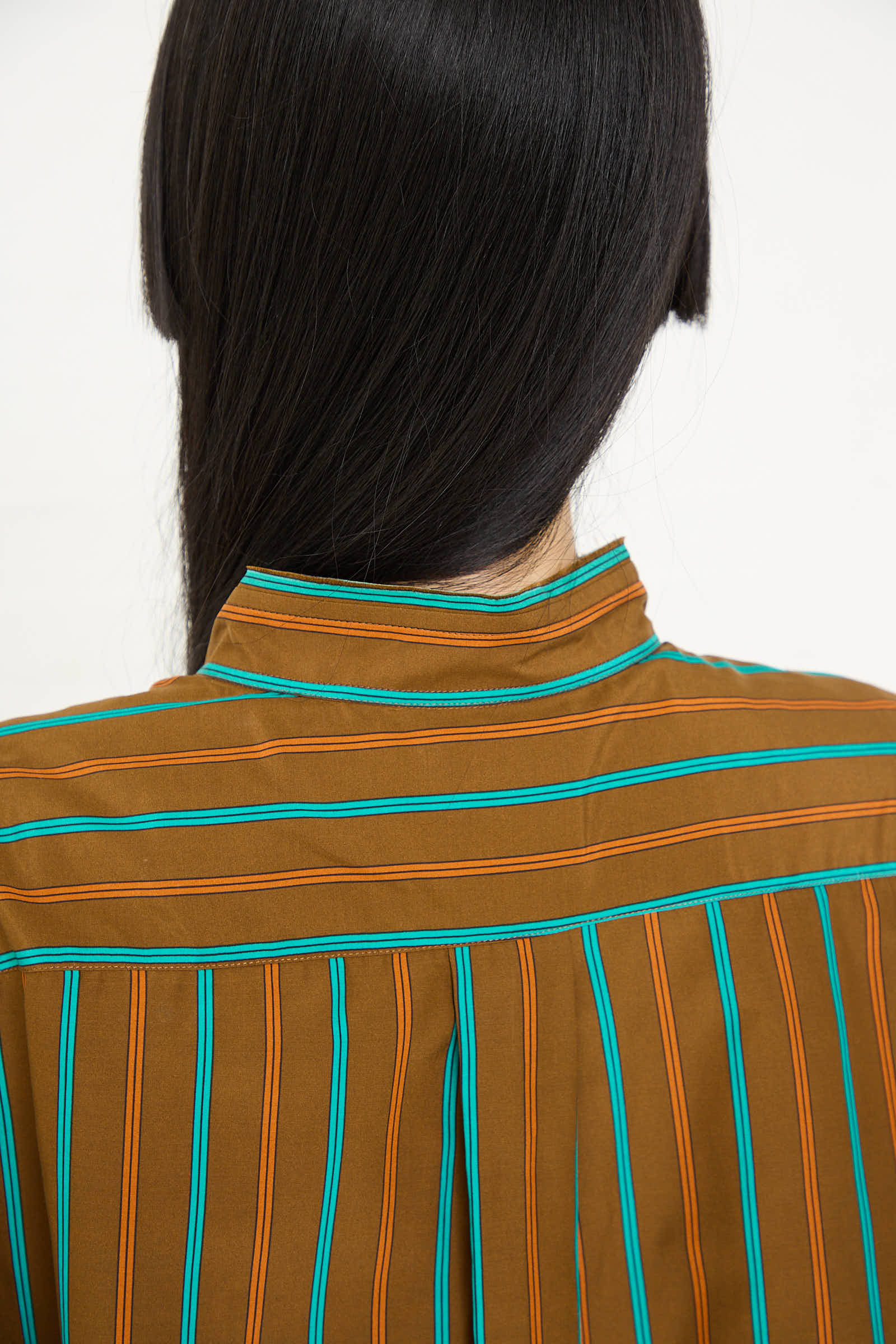 Back view of a person with long black hair wearing a Cristaseya Handmade Fringed Mao Collar Shirt in Noisette with Blue Stripe.