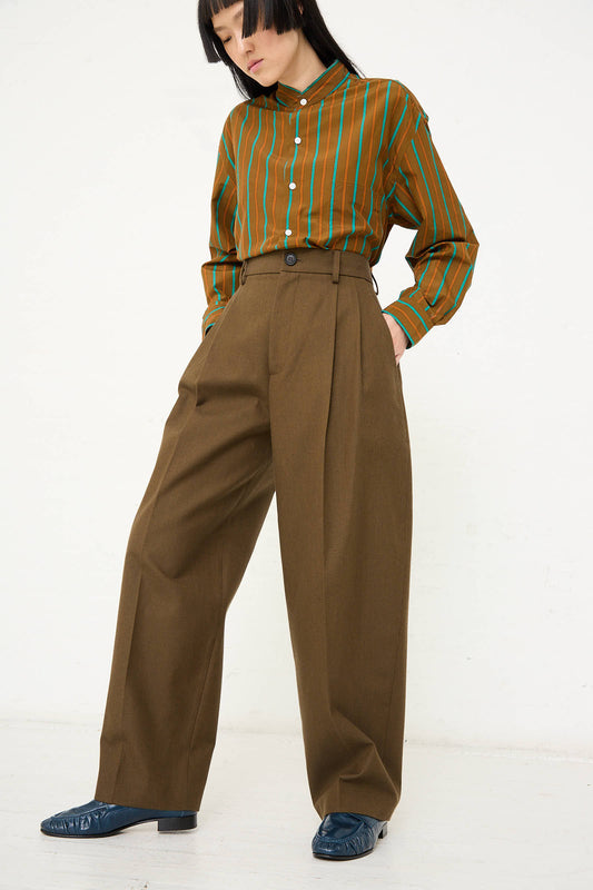 Person standing against a white background, wearing a green and brown striped shirt, Cristaseya's Japanese Wool and Cotton Twill Double Pleated Wide Pant in Dark Olive, and black shoes.