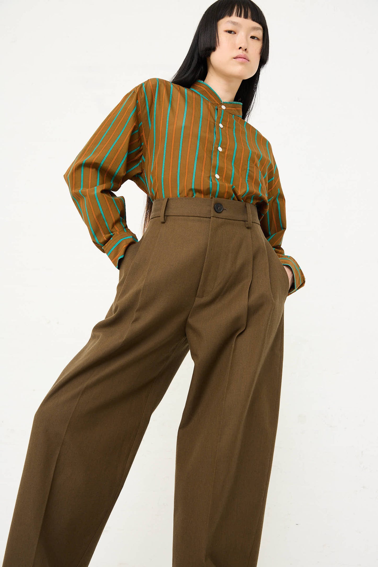 A person wearing a green and brown striped shirt paired with Cristaseya's Japanese Wool and Cotton Twill Double Pleated Wide Pant in Dark Olive is standing against a plain background, looking slightly upward.