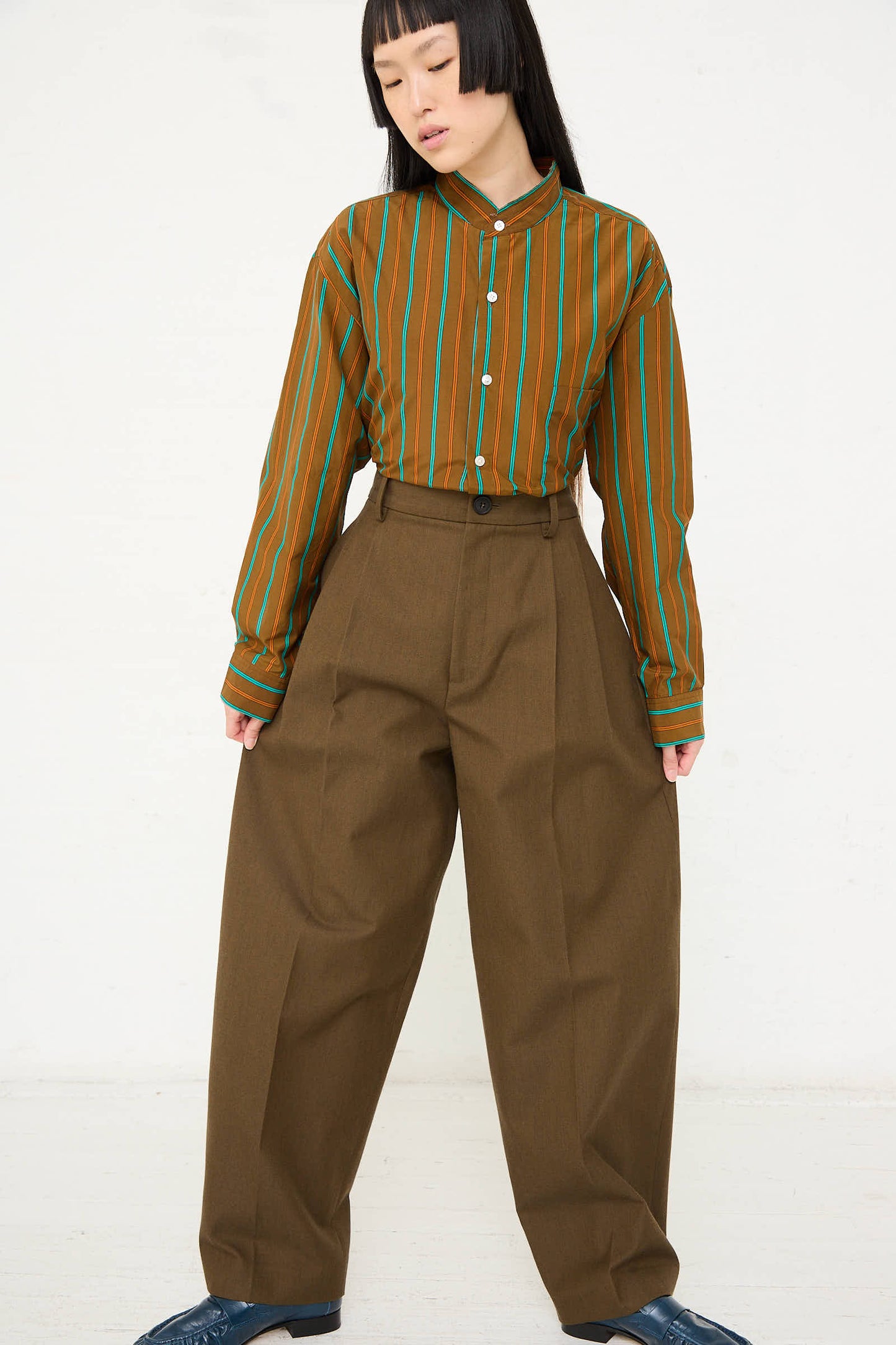 A person stands against a plain background, wearing a green and orange striped shirt, the Cristaseya Japanese Wool and Cotton Twill Double Pleated Wide Pants in Dark Olive, and blue shoes, looking down with hands by their sides.