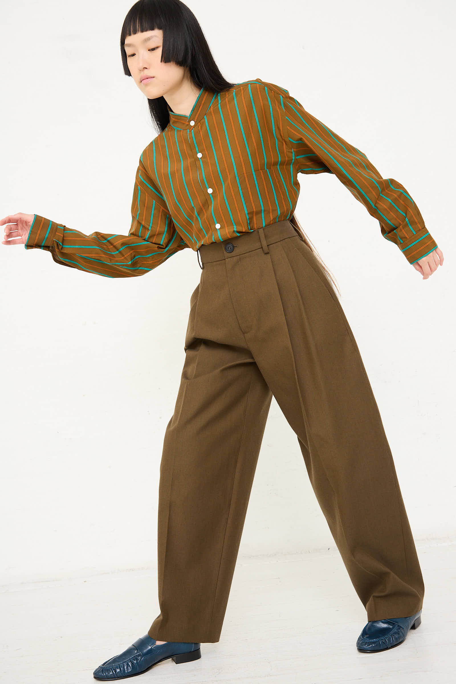 A person wearing a brown and green striped long-sleeve shirt, Cristaseya's Japanese Wool and Cotton Twill Double Pleated Wide Pant in Dark Olive, and blue shoes stands against a plain white background.