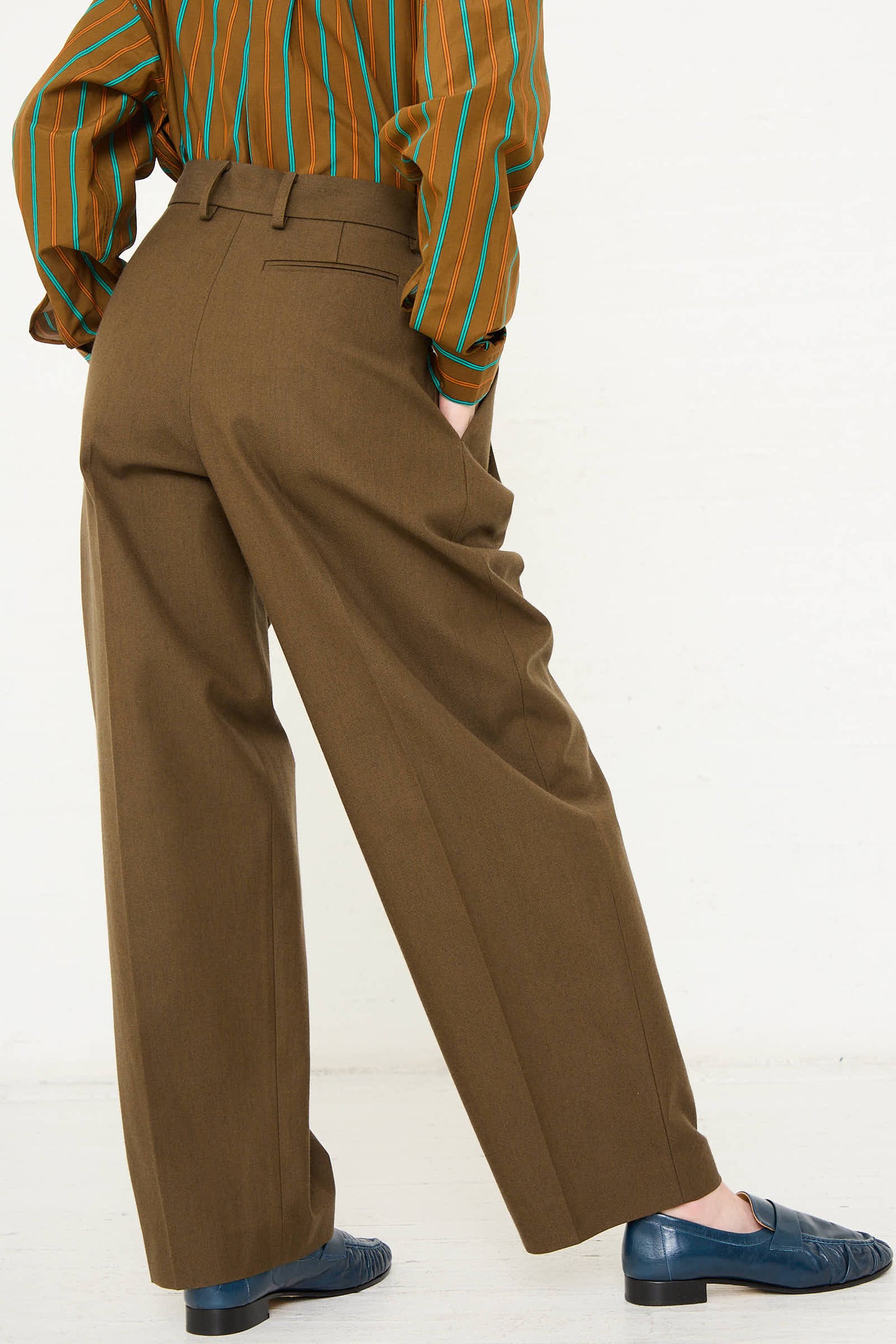 Person wearing dark olive double pleated wide pants made from a Japanese wool and cotton twill blend by Cristaseya, and a brown striped shirt, standing with their back to the camera.