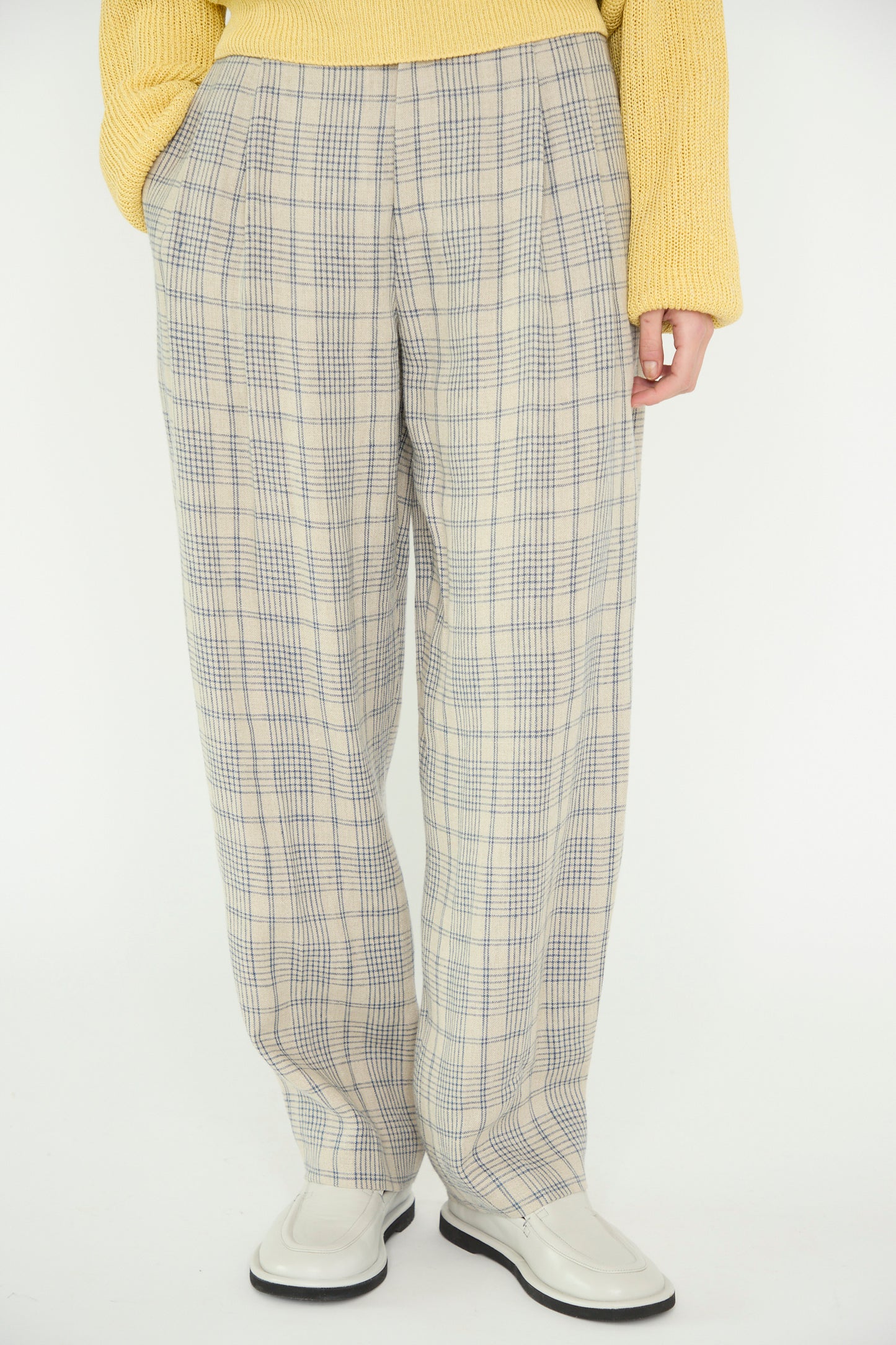 Person wearing Cristaseya's Linen Double Pleated Wide Pant in Cream with Petrol Checks, a yellow sweater, and white shoes standing against a plain background.