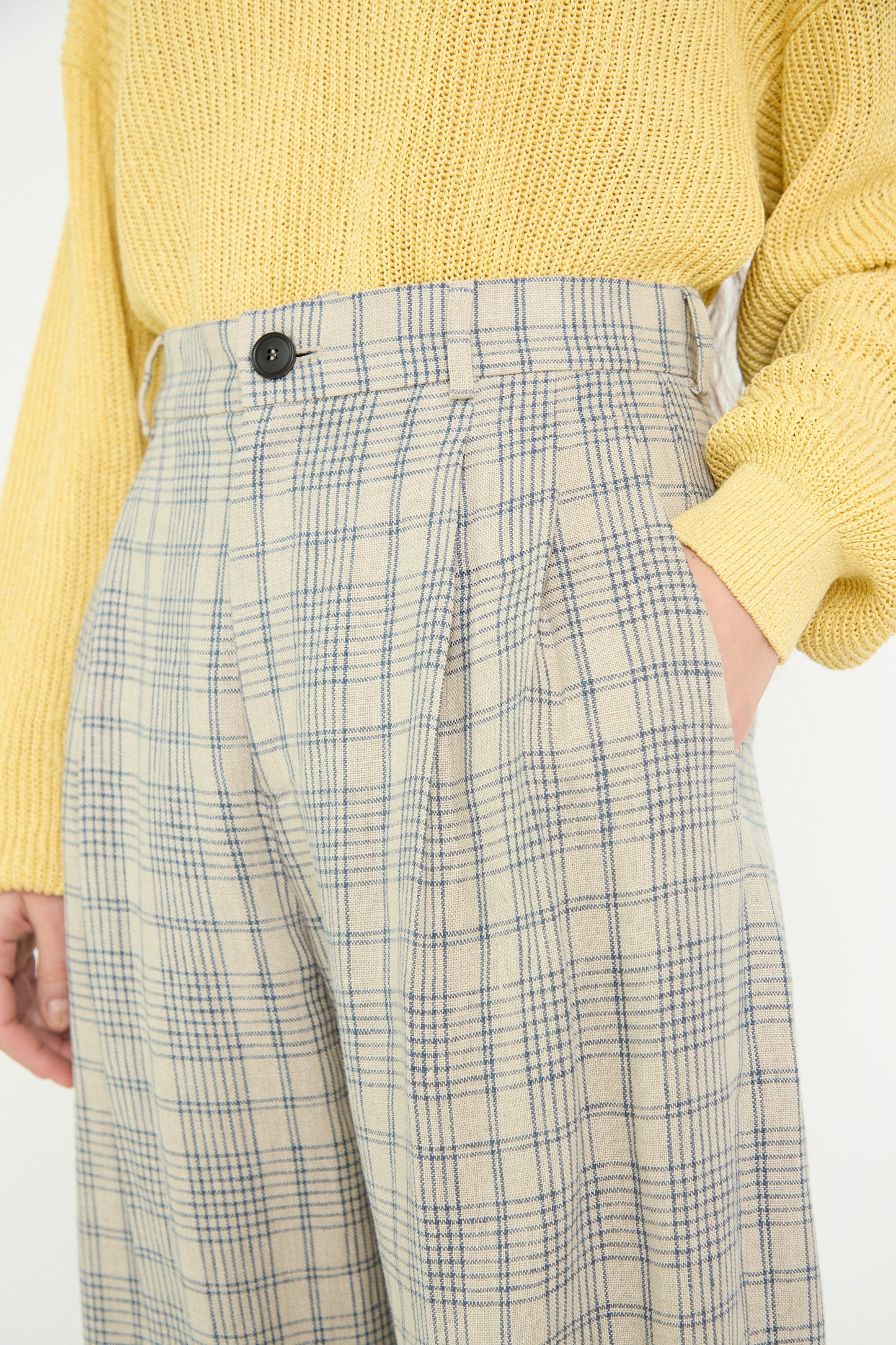 A close-up of a model wearing a yellow sweater with Cristaseya's Linen Double Pleated Wide Pant in Cream with Petrol Checks, hand in pockets.